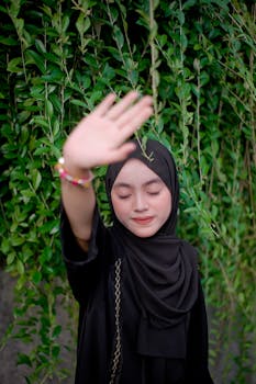 A young woman wearing a black hijab stands amidst green foliage, enjoying nature.