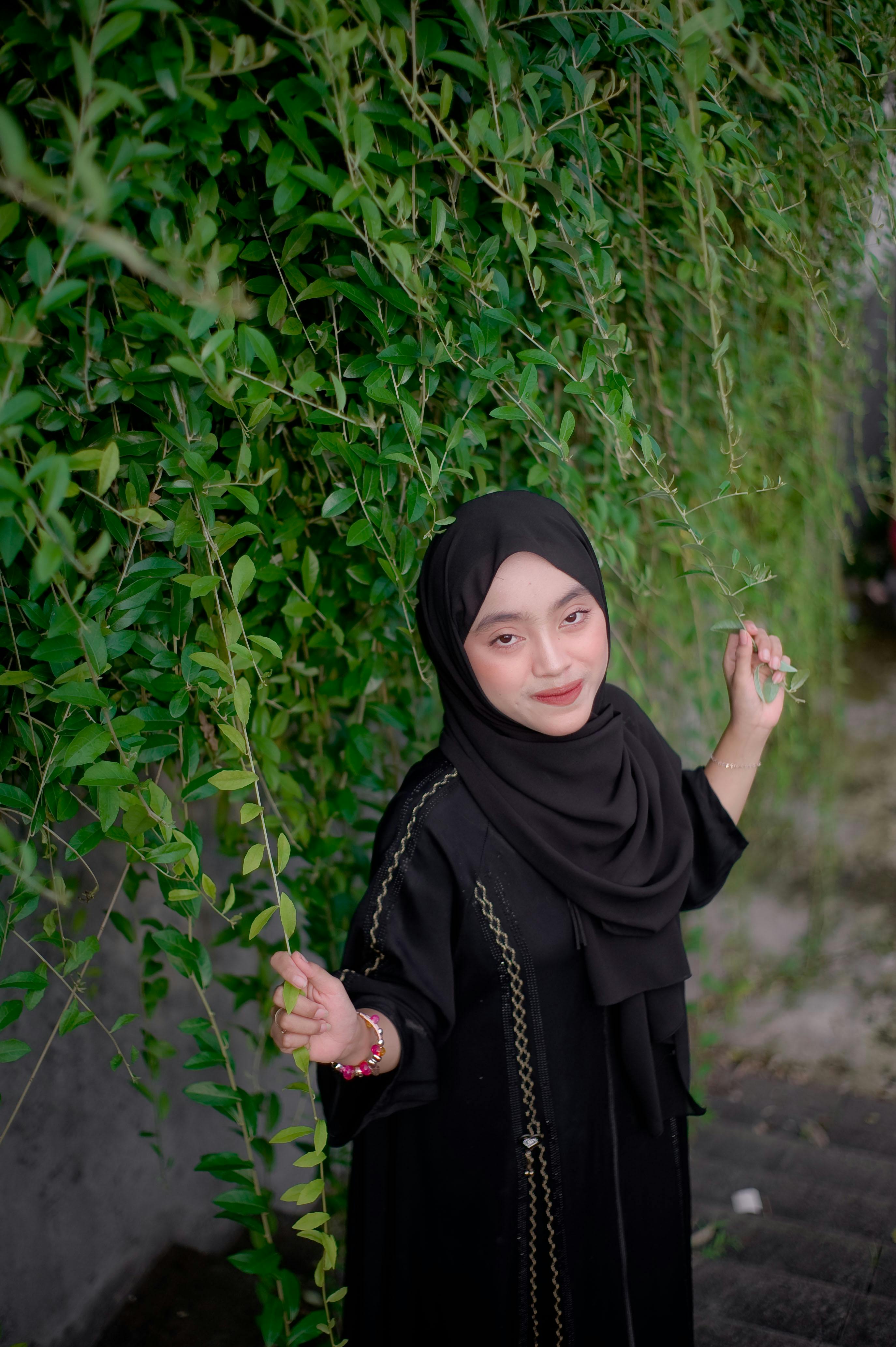 Woman in black hijab smiling next to lush green foliage outdoors.