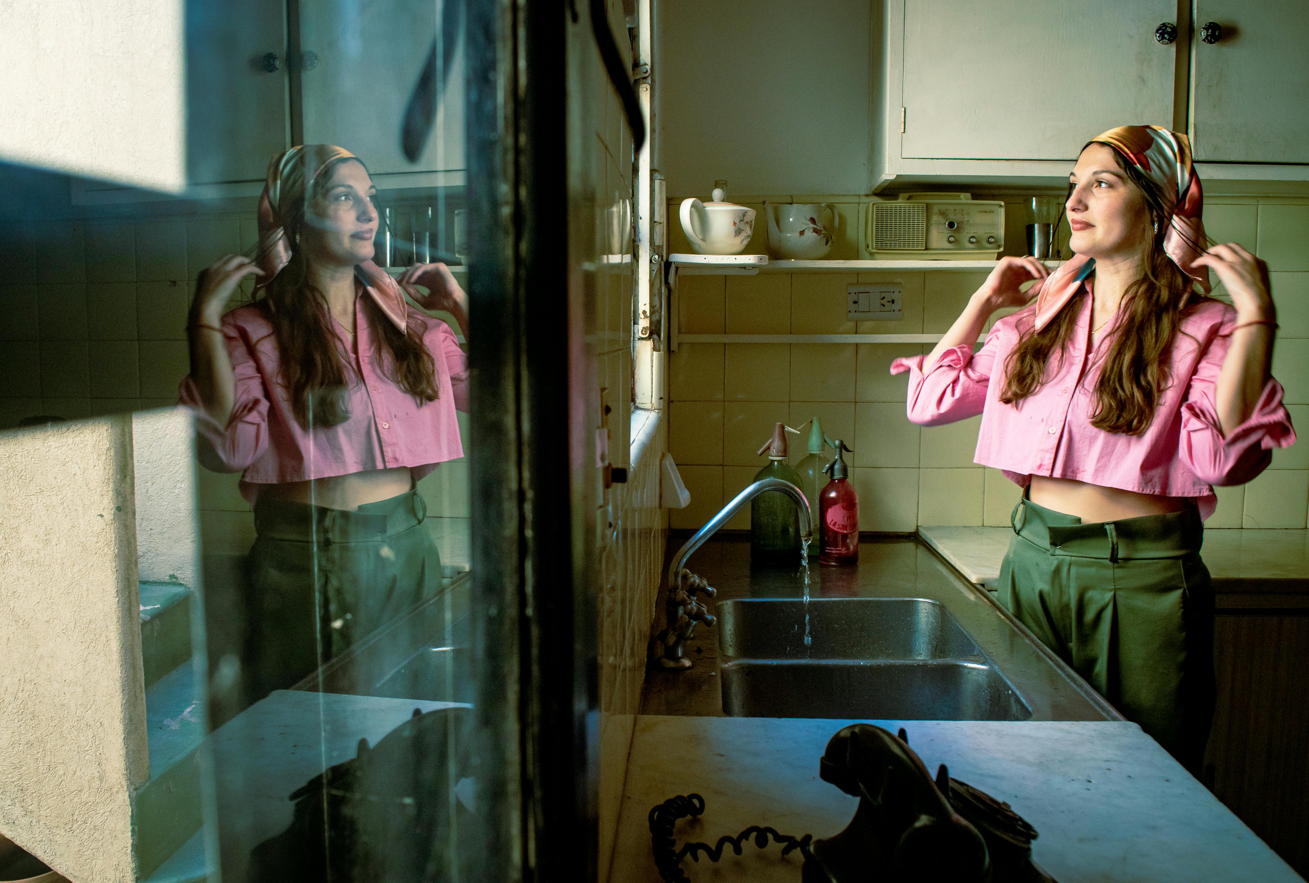 Ücretsiz Eski tarz bir mutfakta, pencerede yansıyan görüntüsü eşliğinde bir kadın başörtüsünü düzeltiyor. Canlı ve nostaljik bir atmosfer. Stok Fotoğraflar