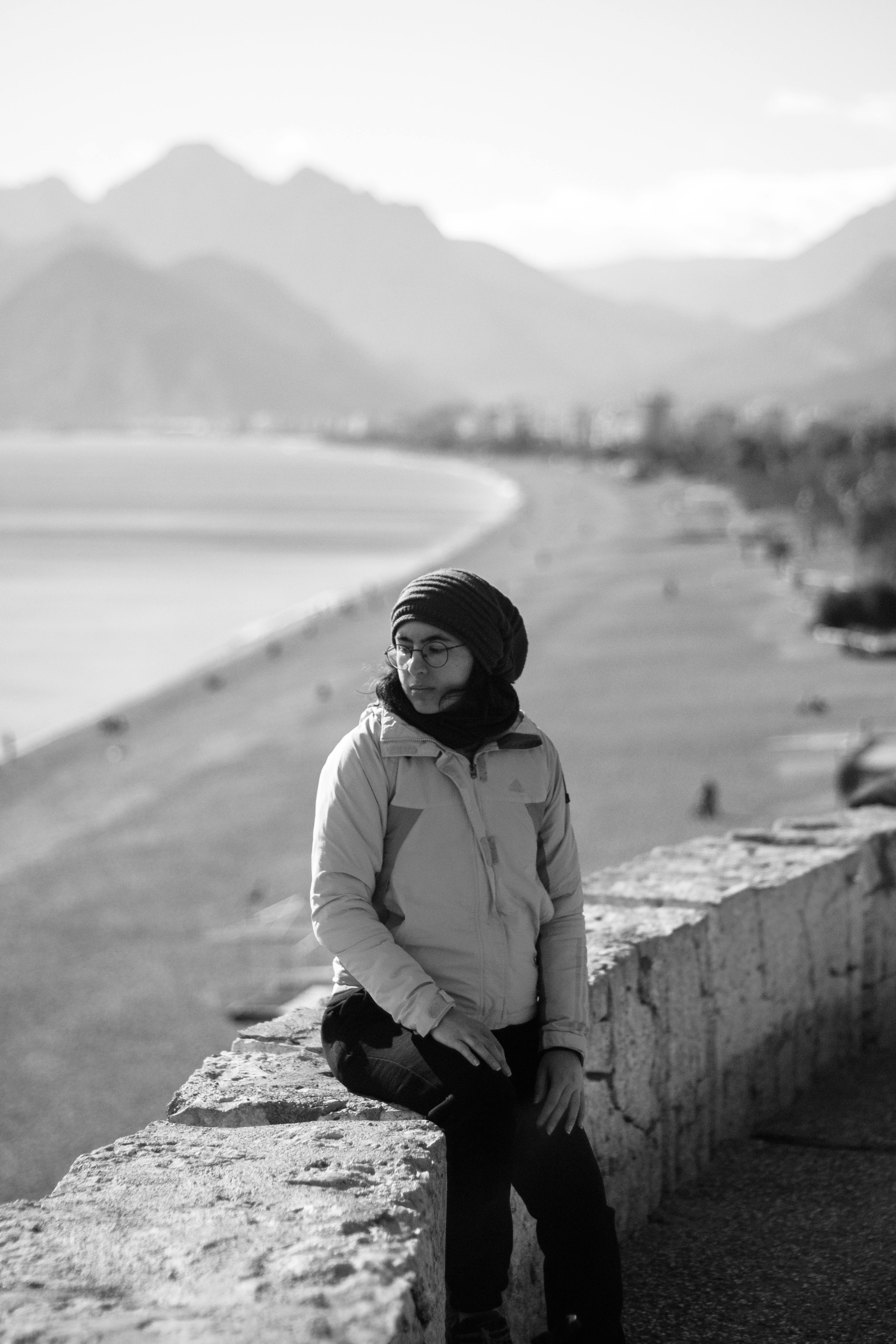 Ücretsiz Siyah beyaz bir fotoğrafta, manzarası muhteşem dağlık bir kıyı şeridindeki taş bir duvarın üzerinde oturan bir kadın. Stok Fotoğraflar