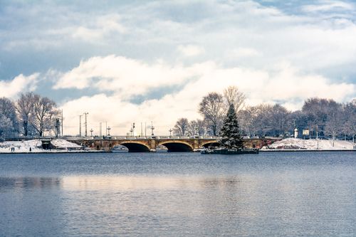 Free Scenic winter view of a bridge over a calm lake surrounded by snow-covered trees and a cloudy sky. Stock Photo