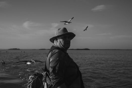 Black and white photo of a man in a hat near the sea with flying birds.