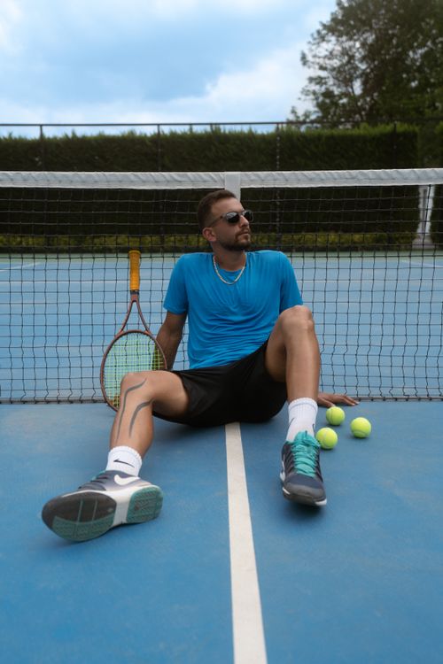 Free A young man in sportswear relaxes against a tennis net on an outdoor court, with tennis balls and racket. Stock Photo