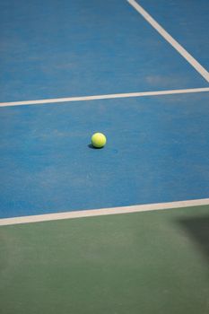 A lone tennis ball rests on a vibrant blue and green court under bright daylight.