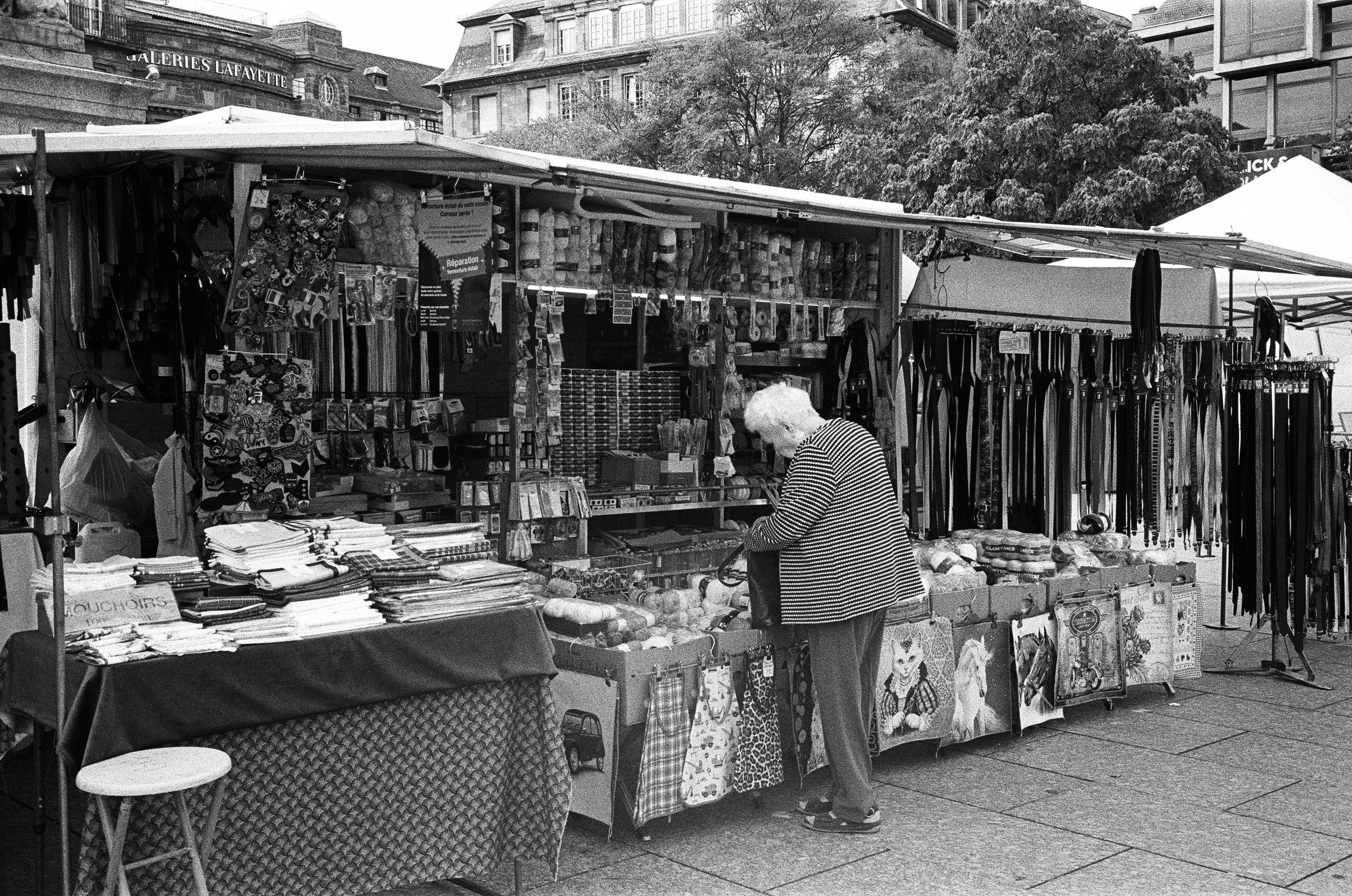 Black and white photo of an elderly woman exploring a bustling outdoor market in Strasbourg.