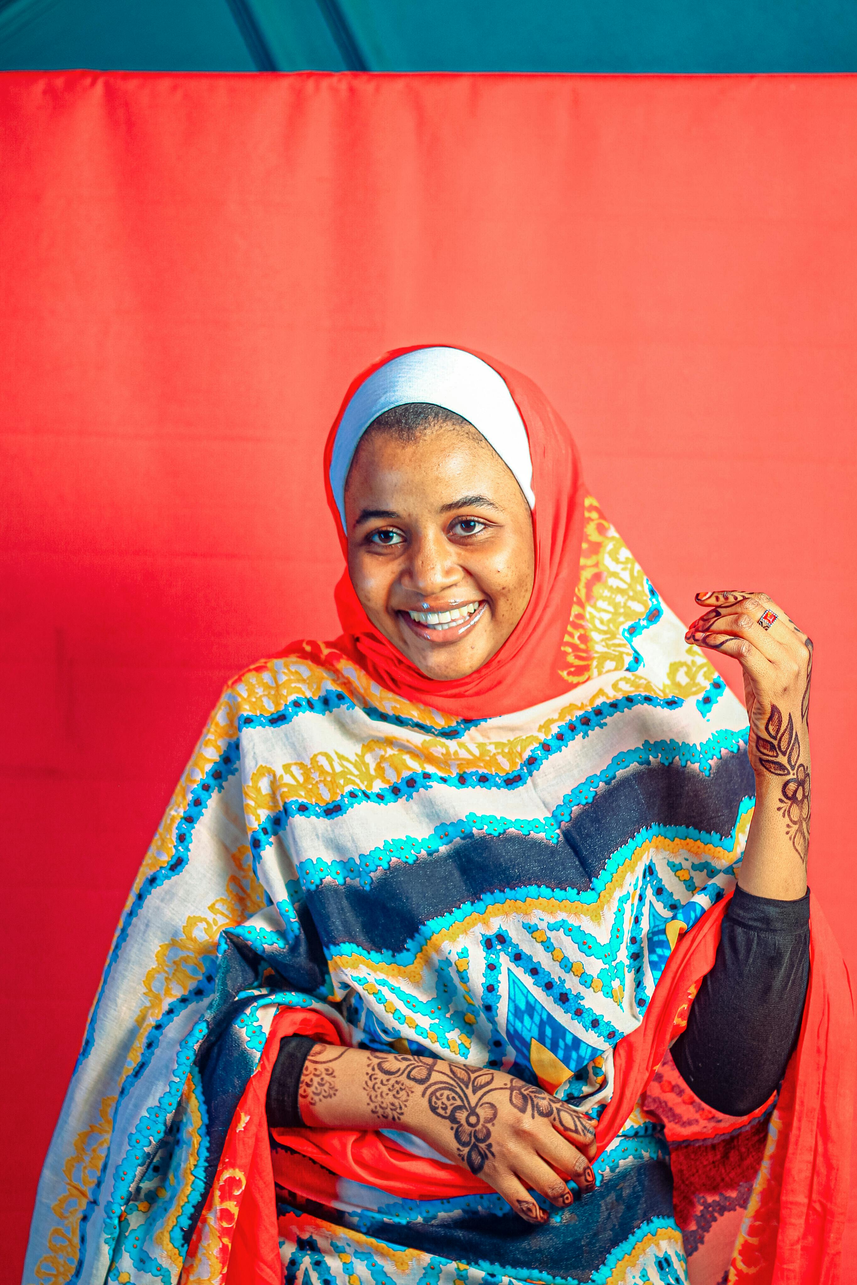 A cheerful woman wearing a colorful hijab and intricate henna designs, set against a bright background.