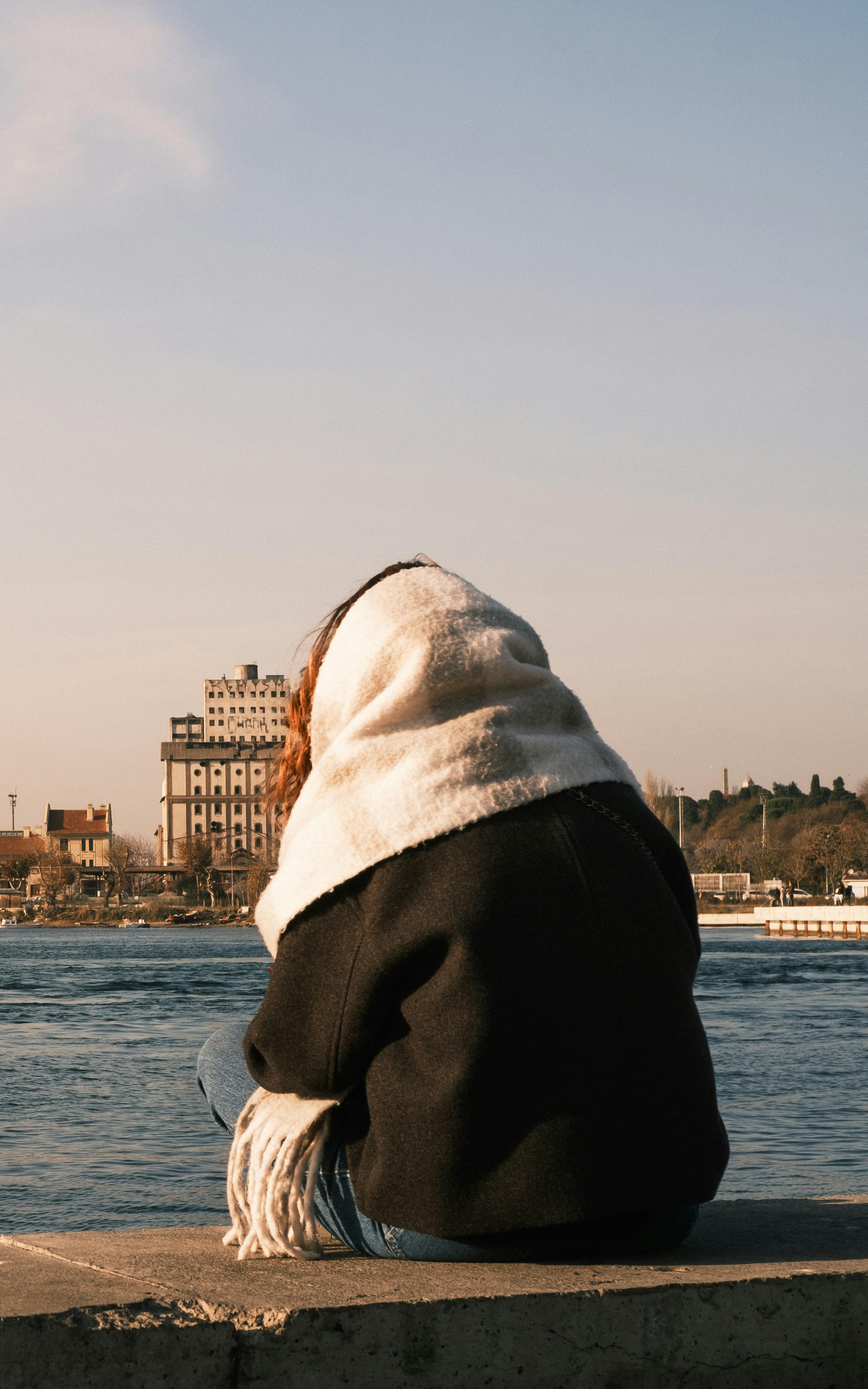Bezpłatne Kobieta w szaliku siedzi zwrócona twarzą w stronę spokojnej rzeki podczas zachodu słońca w miejskim krajobrazie. Zdjęcie z galerii