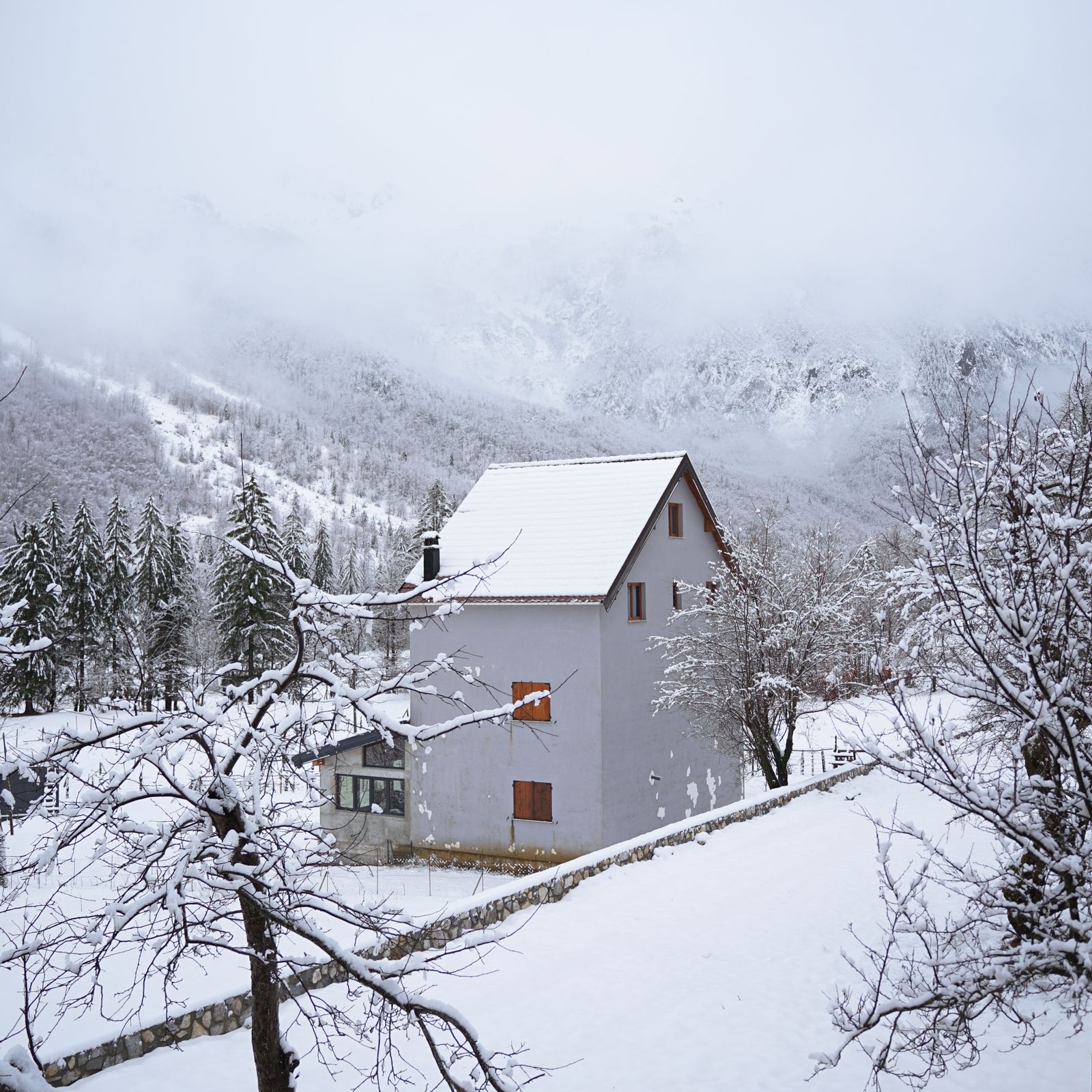Maison enneigée et sapins sous des sommets brumeux et enneigés à Valbonë, Albanie