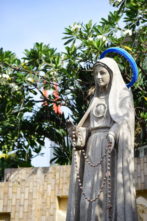 Free A stone statue of a religious figure holding a rosary in a garden setting with trees. Stock Photo