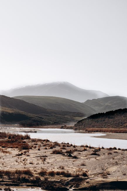 Serene mountain landscape featuring a calm river and rolling hills under a bright sky.
