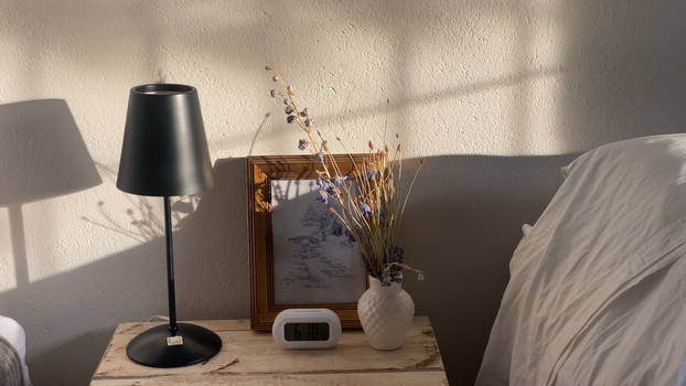 Sunlit bedroom interior with a lamp, vase, and clock on a wooden table, creating a cozy atmosphere.
