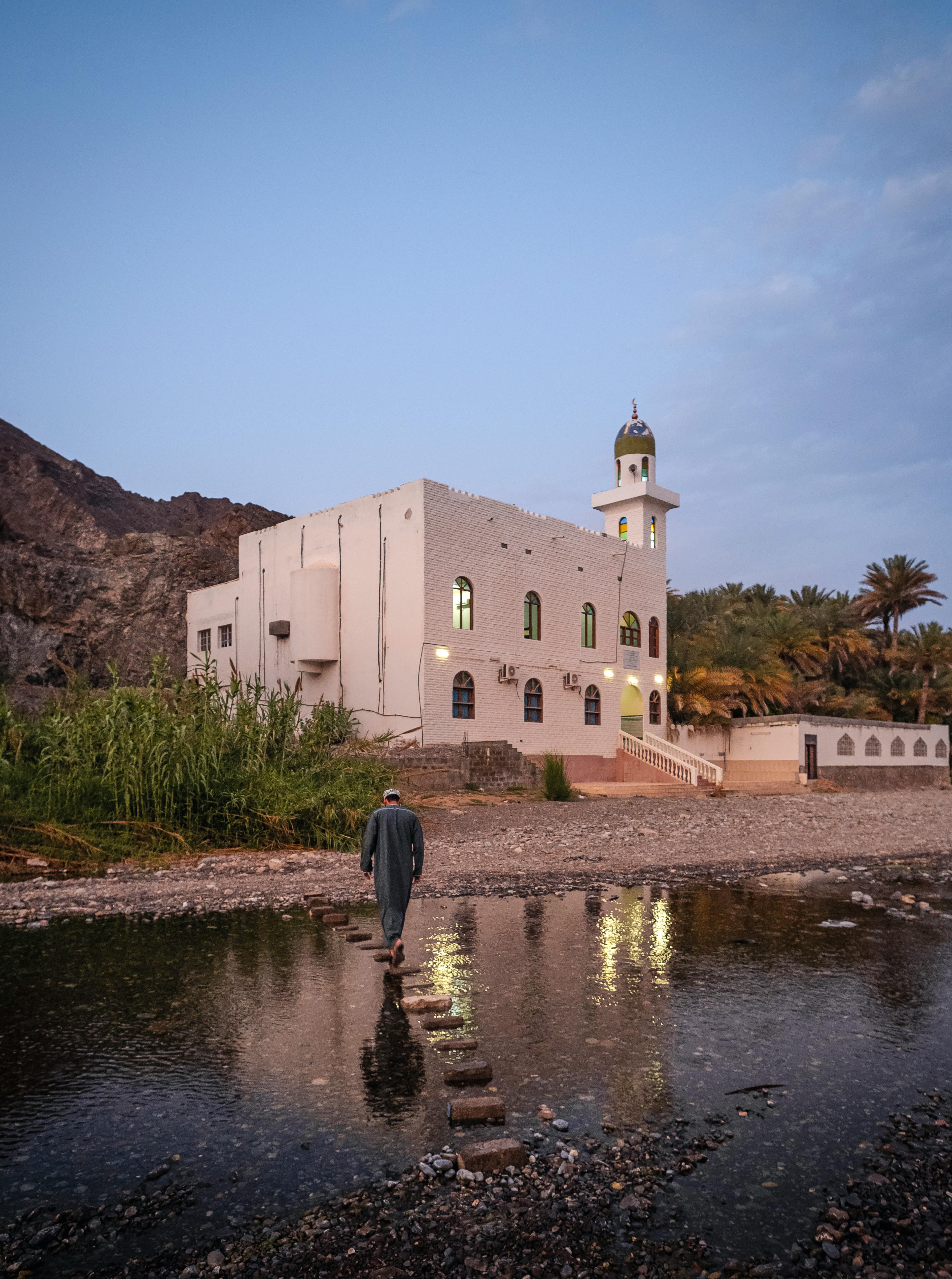 Man crossing stepping stones towards a mosque during twilight in Oman, with a tranquil setting and surrounding nature.