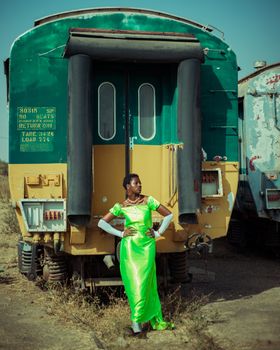 Stylish woman poses in a bright green dress in front of a vintage train under clear skies.