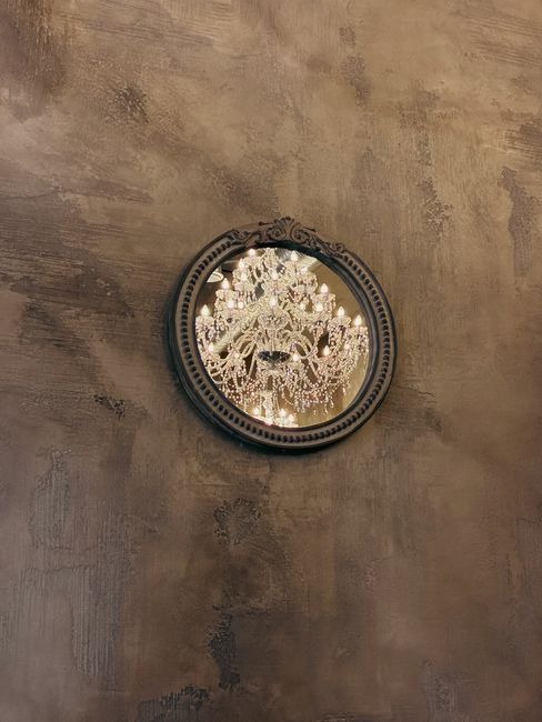 Reflection of a crystal chandelier in a decorative, round mirror on a textured wall.