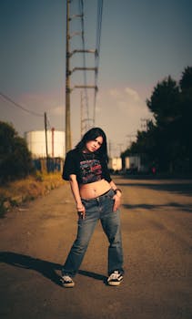 Young adult posing in casual urban fashion amidst industrial backdrop during sunset.