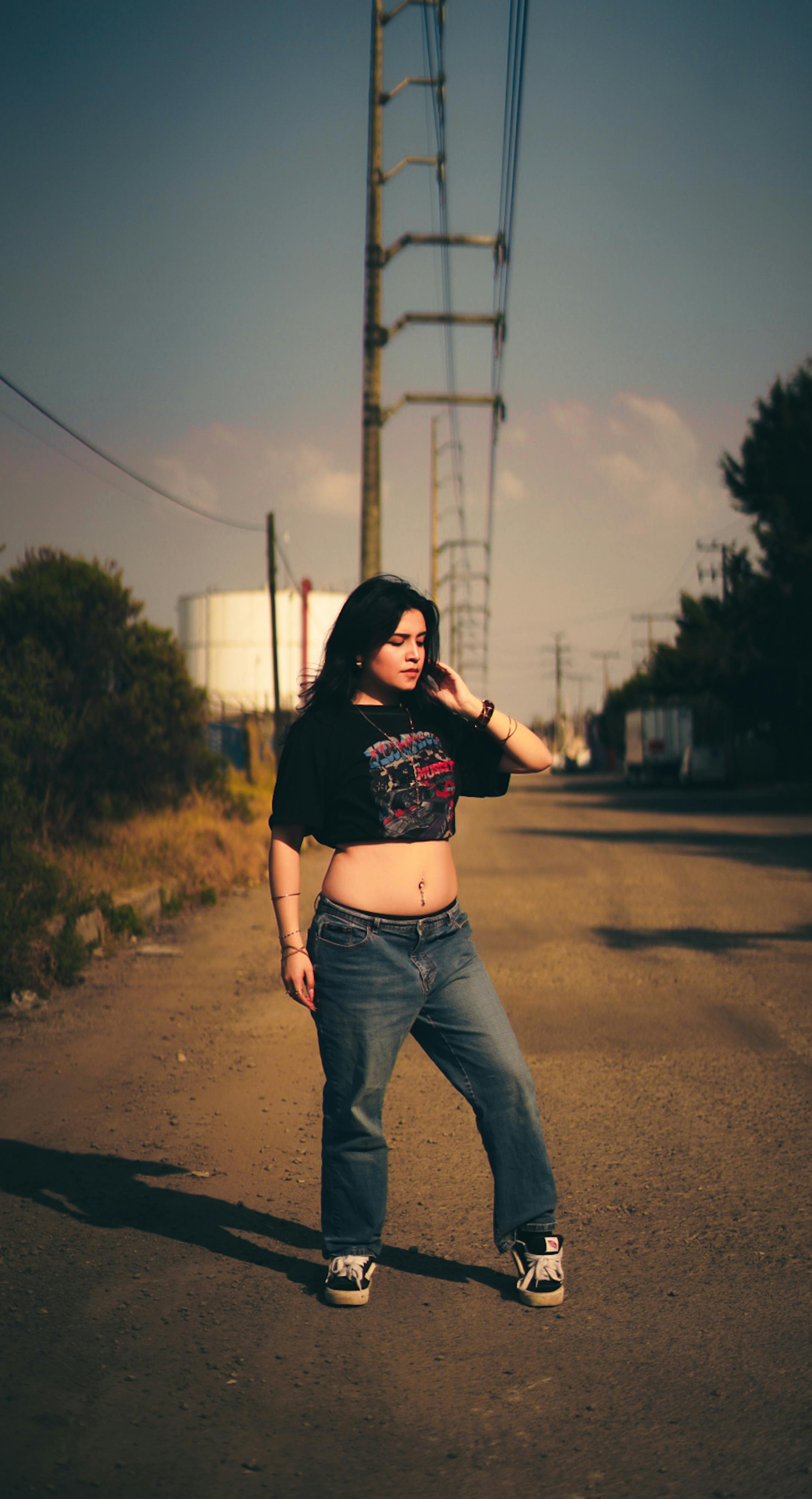 Gratis Seorang wanita muda yang percaya diri berpose di jalan tanah pedesaan di bawah kabel telepon. Foto Stok