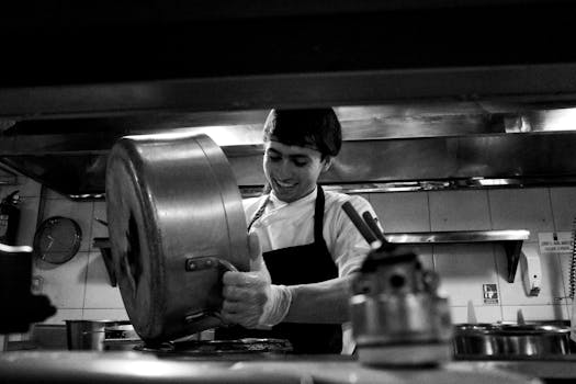A chef skillfully pours from a large saucepan in a bustling restaurant kitchen.