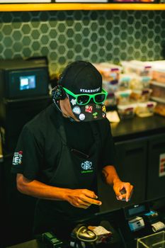 Barista wearing hat and sunglasses working at a coffee shop counter with pastry display in background.
