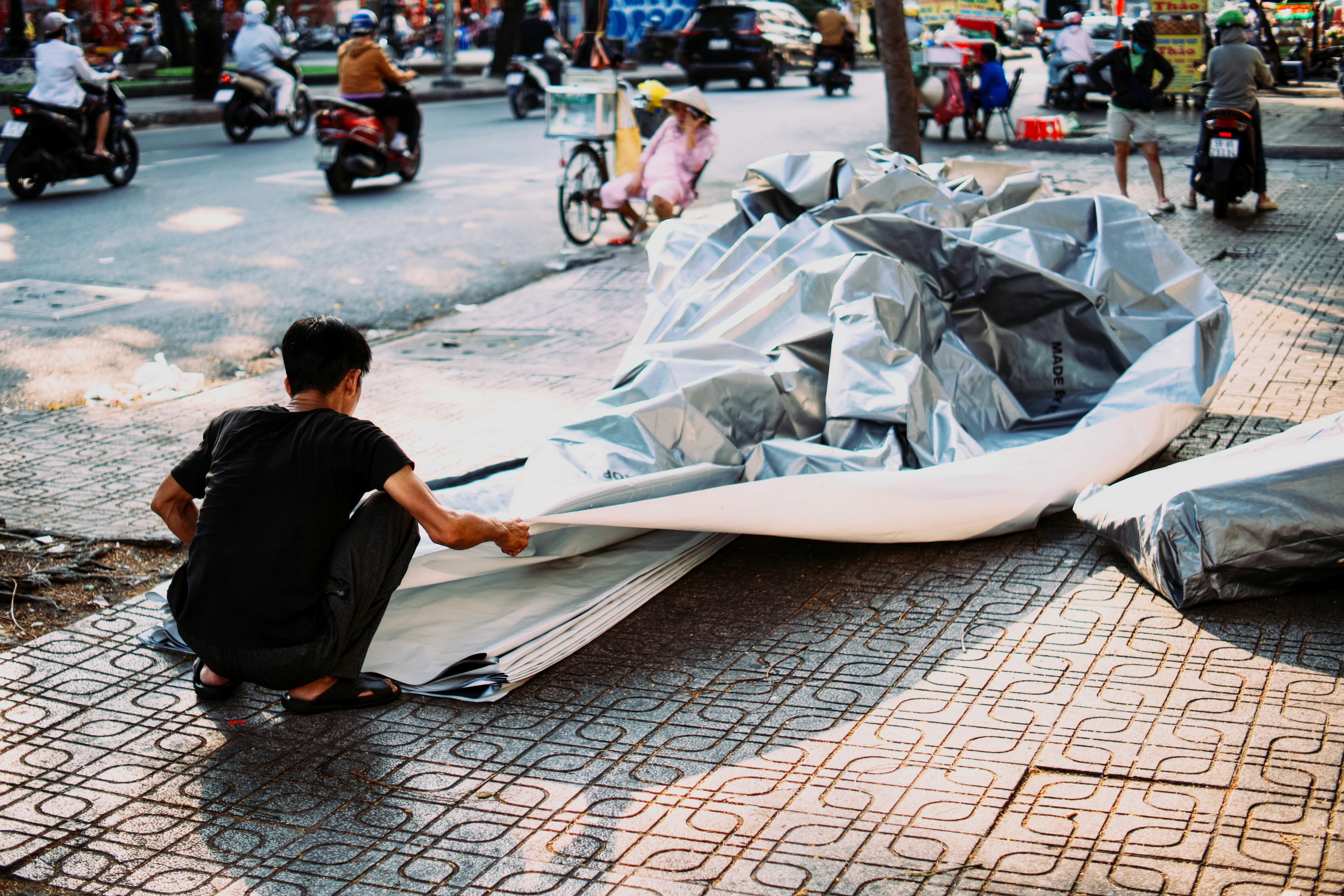 Gratis Seorang pria membongkar barang-barang di trotoar perkotaan yang ramai sementara orang-orang lewat dengan skuter. Foto Stok