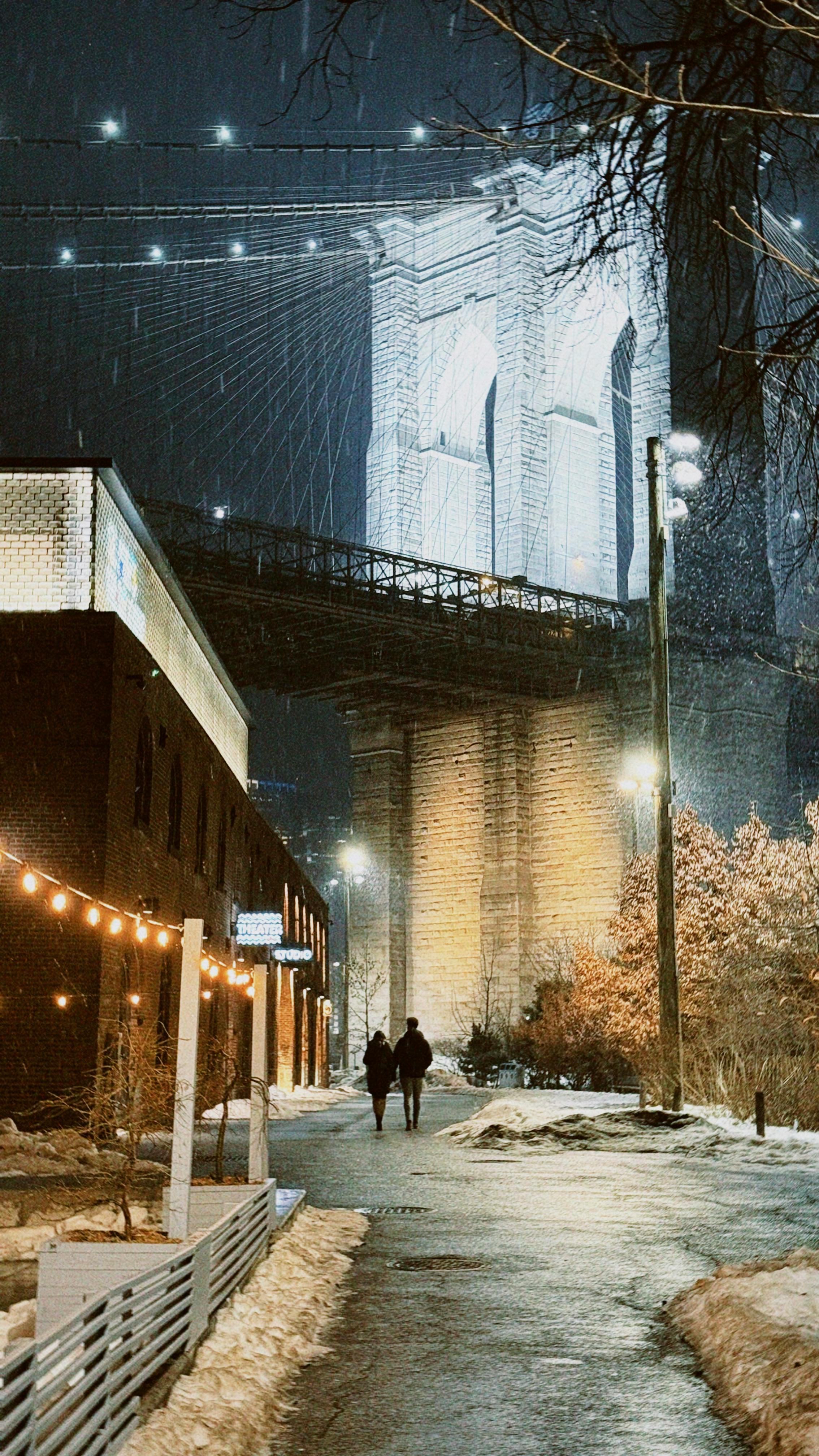 Gratis Sepasang kekasih berjalan di bawah Jembatan Brooklyn yang diterangi cahaya di malam hari, dikelilingi salju dan lampu-lampu meriah. Foto Stok