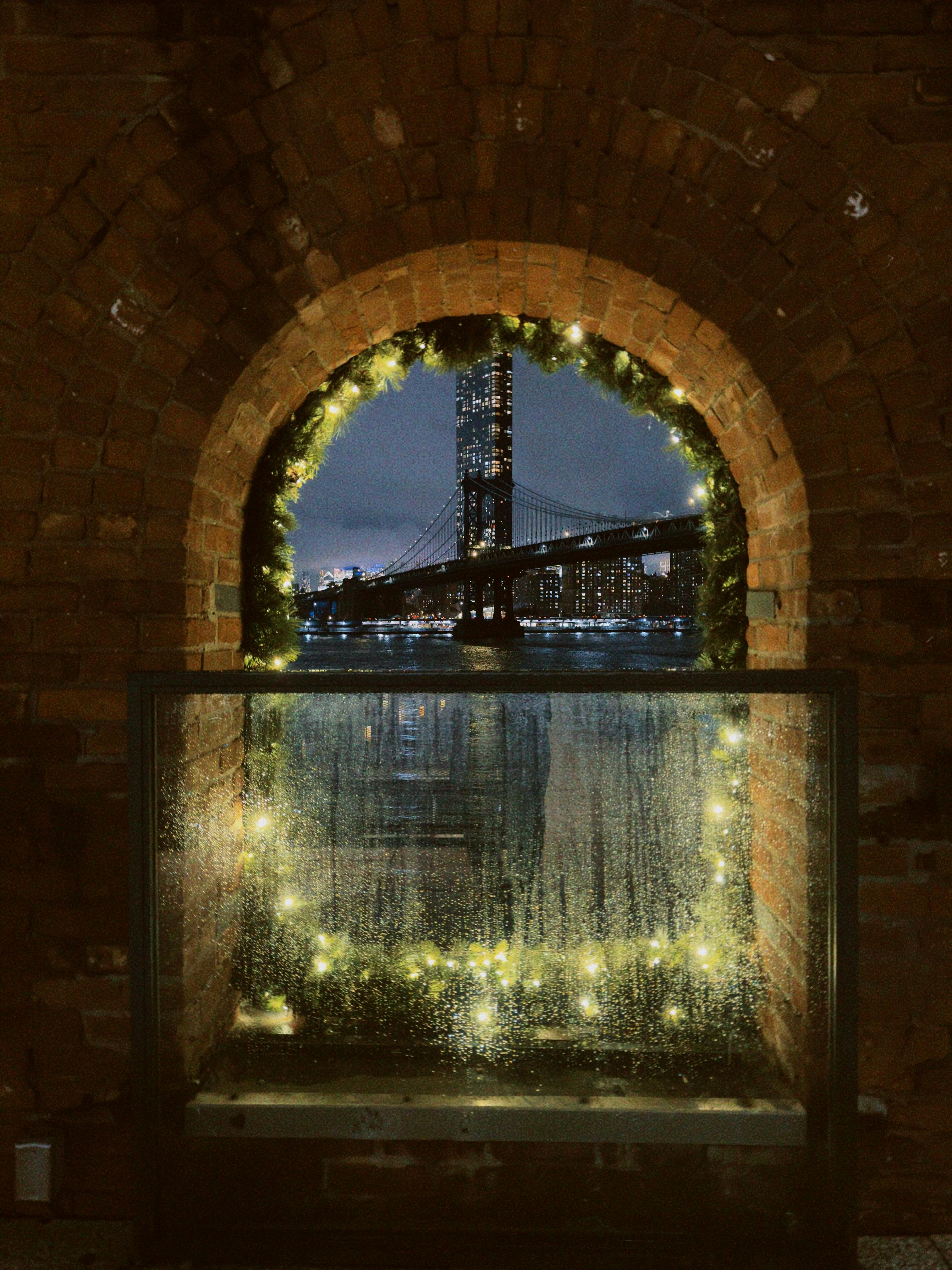 Gratis Pemandangan malam sebuah jembatan yang diterangi lampu melalui gapura bata meriah dengan lampu-lampu peri. Foto Stok