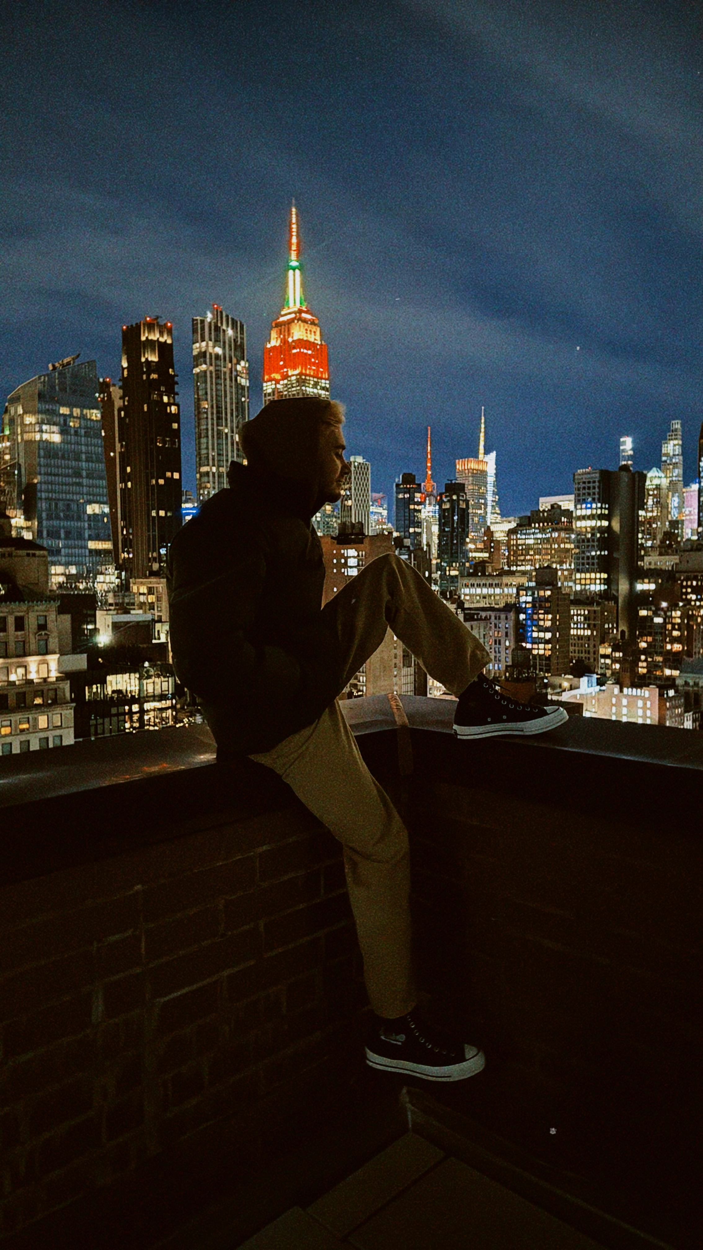 Gratis Siluet seseorang yang duduk di atas atap, memandang cakrawala Kota New York yang semarak di bawah langit malam. Foto Stok