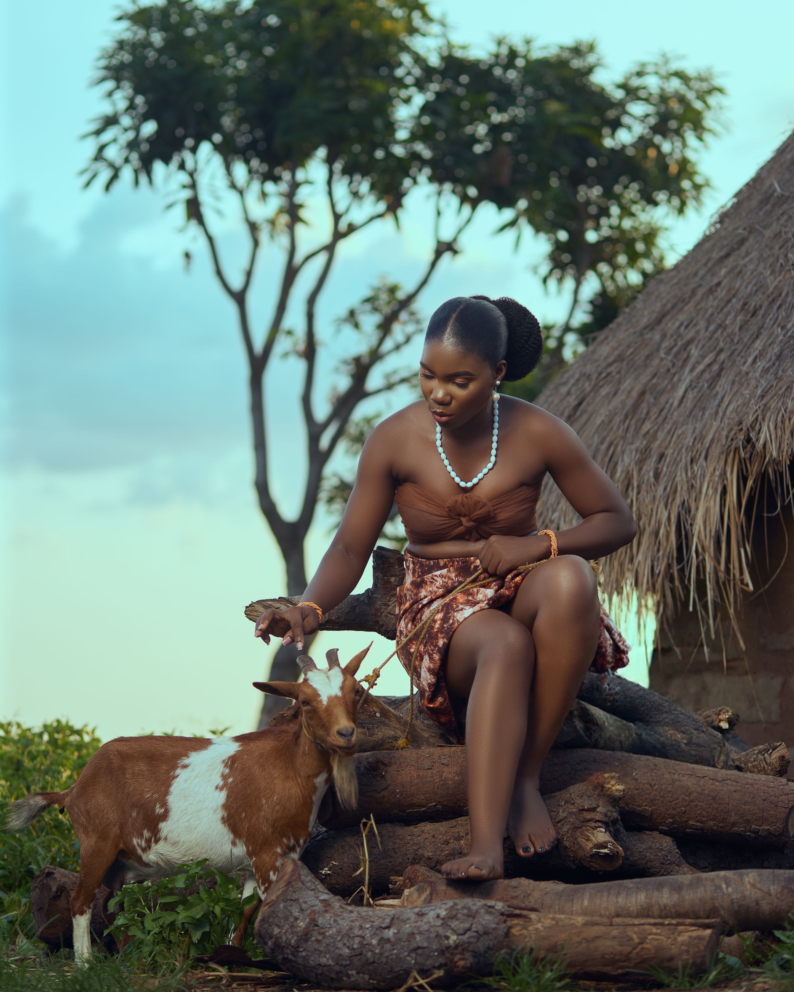 African woman in traditional attire sitting outdoors with a goat, showcasing cultural harmony.