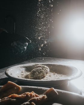 A hand dusting flour over dough, illuminated dramatically in a dark kitchen environment.