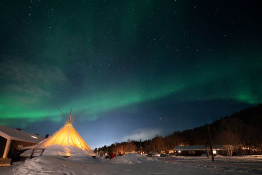 Stunning aurora borealis over a snowy landscape with a lit teepee near Alta, Norway.