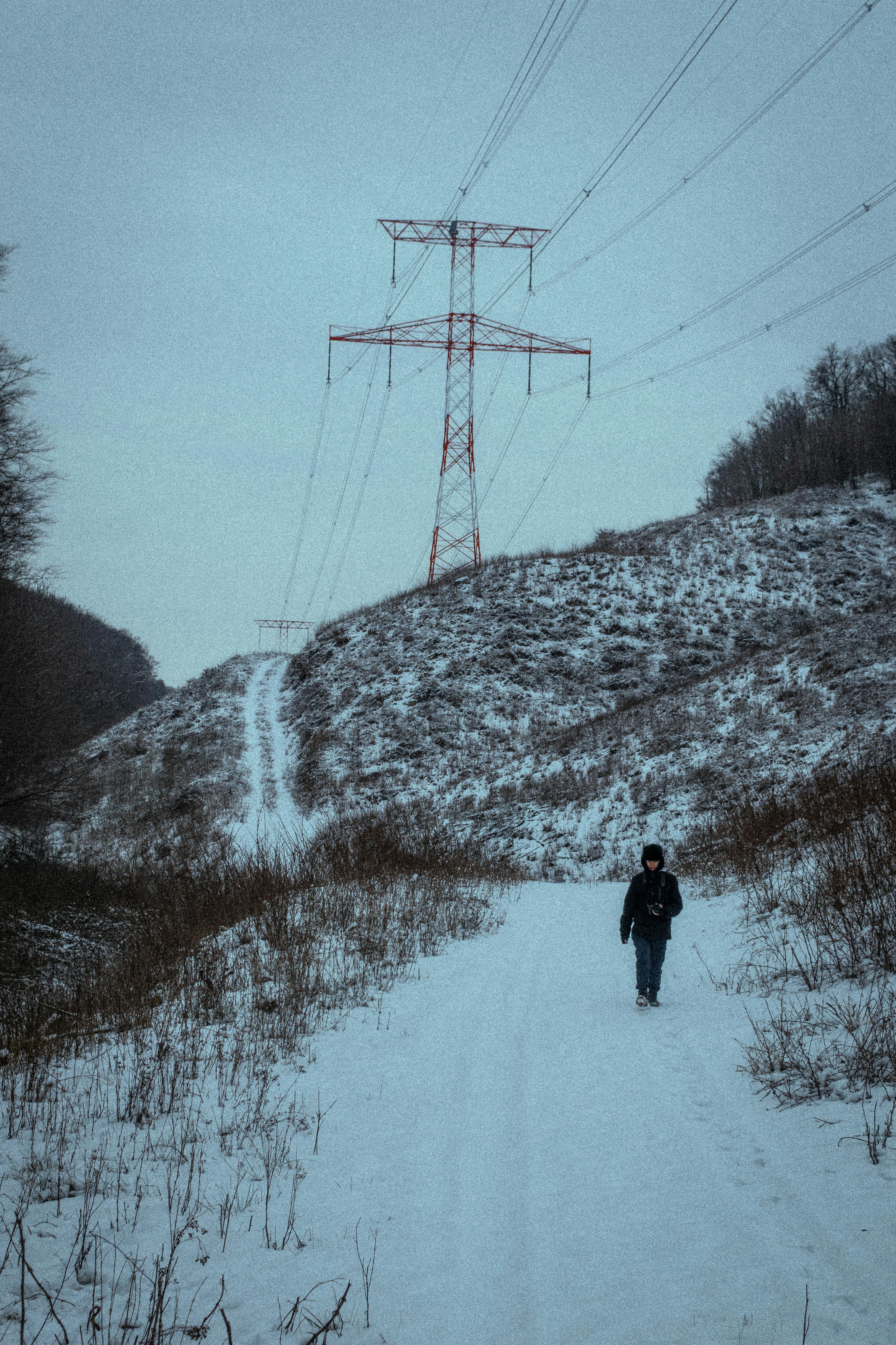 Kostenlos Eine einsame Gestalt wandert durch die verschneite Landschaft unter hoch aufragenden Stromleitungen. Stock-Foto