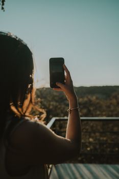 A woman capturing the scenic beauty of sunset using her smartphone, creating a stunning silhouette.