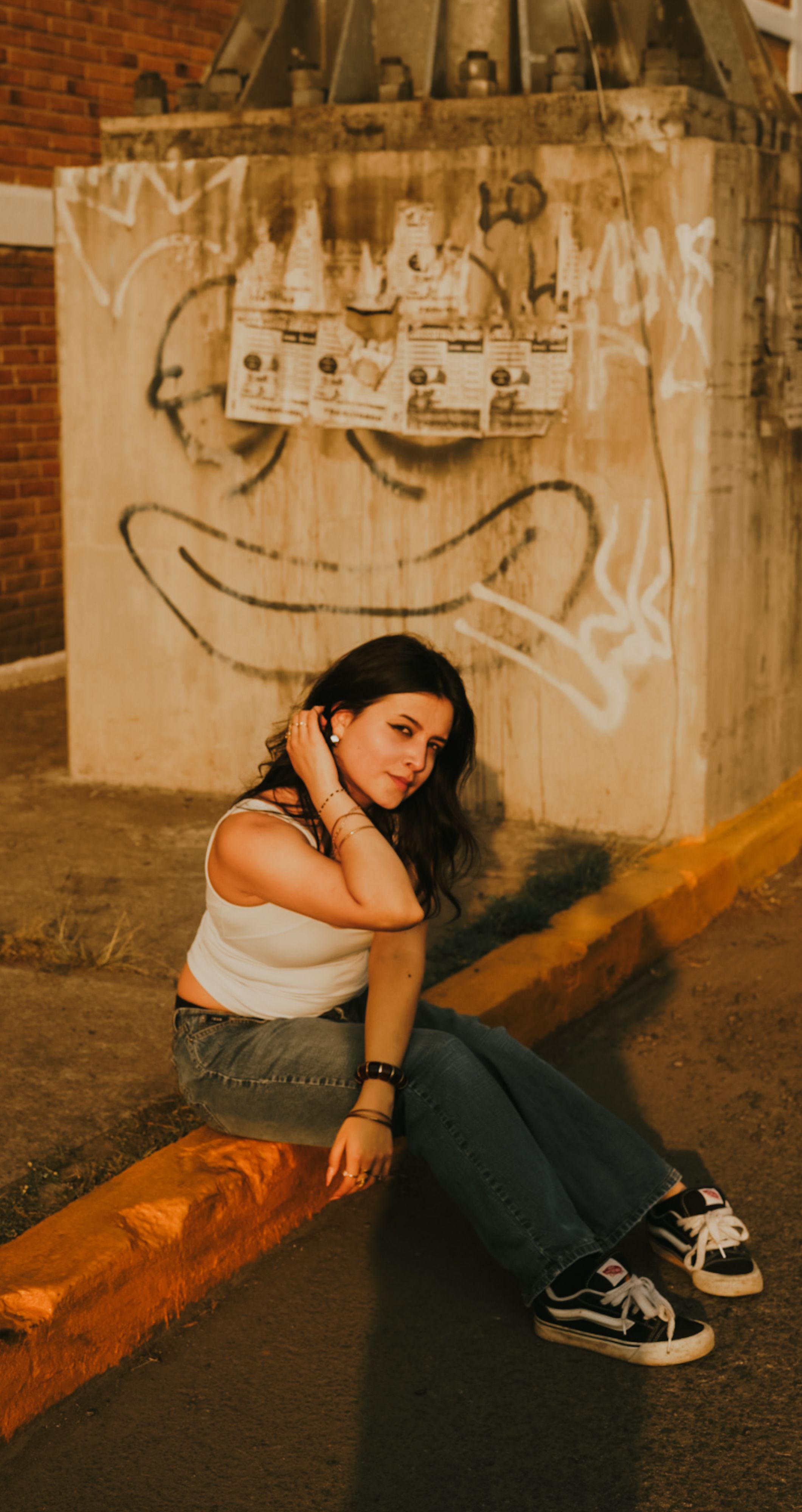Casual portrait of a woman sitting on a curb in an urban setting, with graffiti art on the wall.
