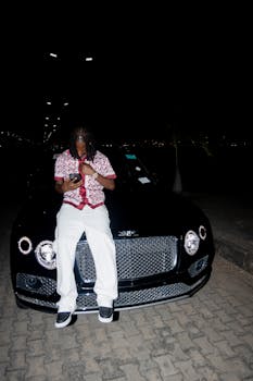 A fashionable man using a smartphone while sitting on a black luxury car at night.