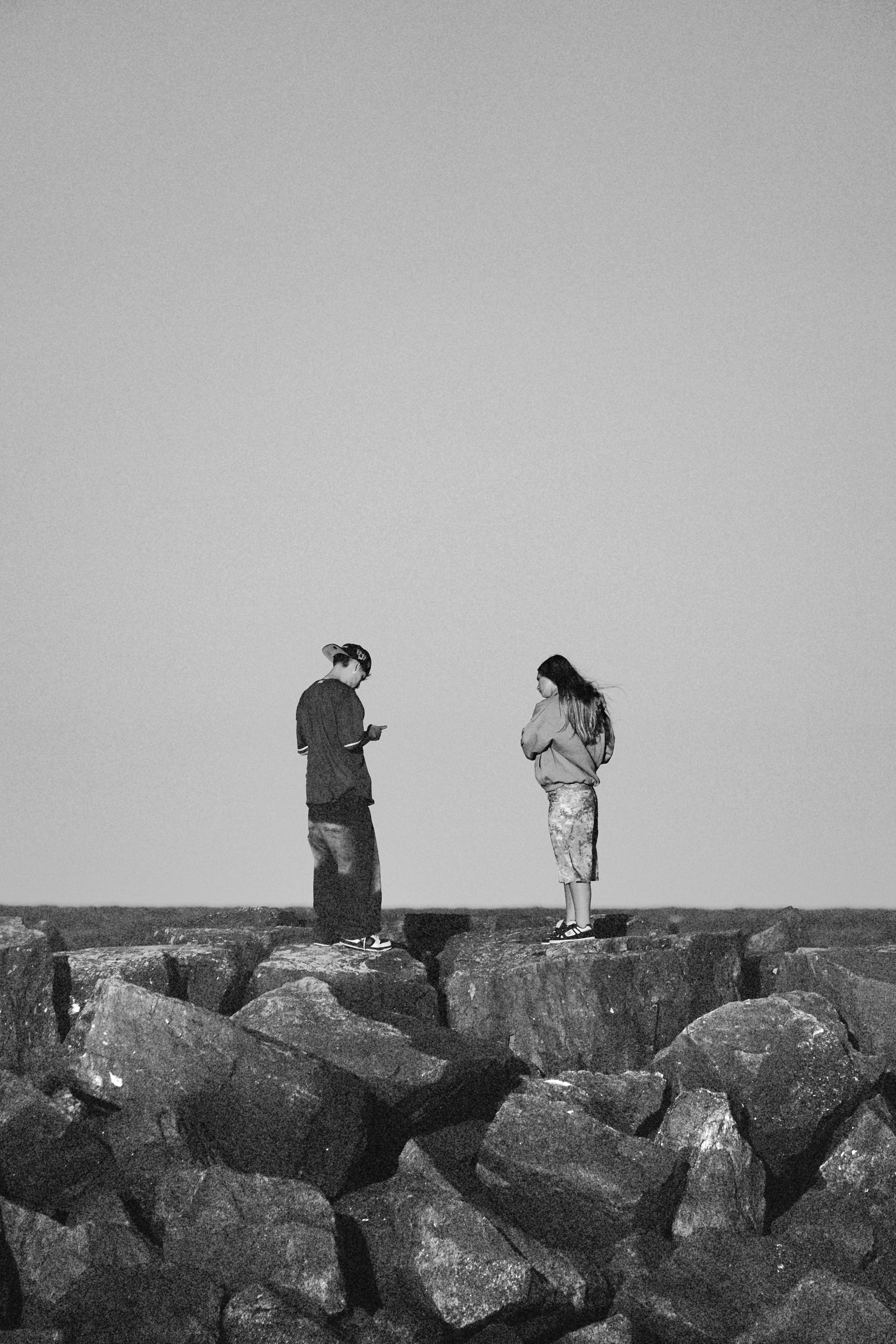 Free Two individuals stand on large rocks by the shore in a black and white scene. Stock Photo