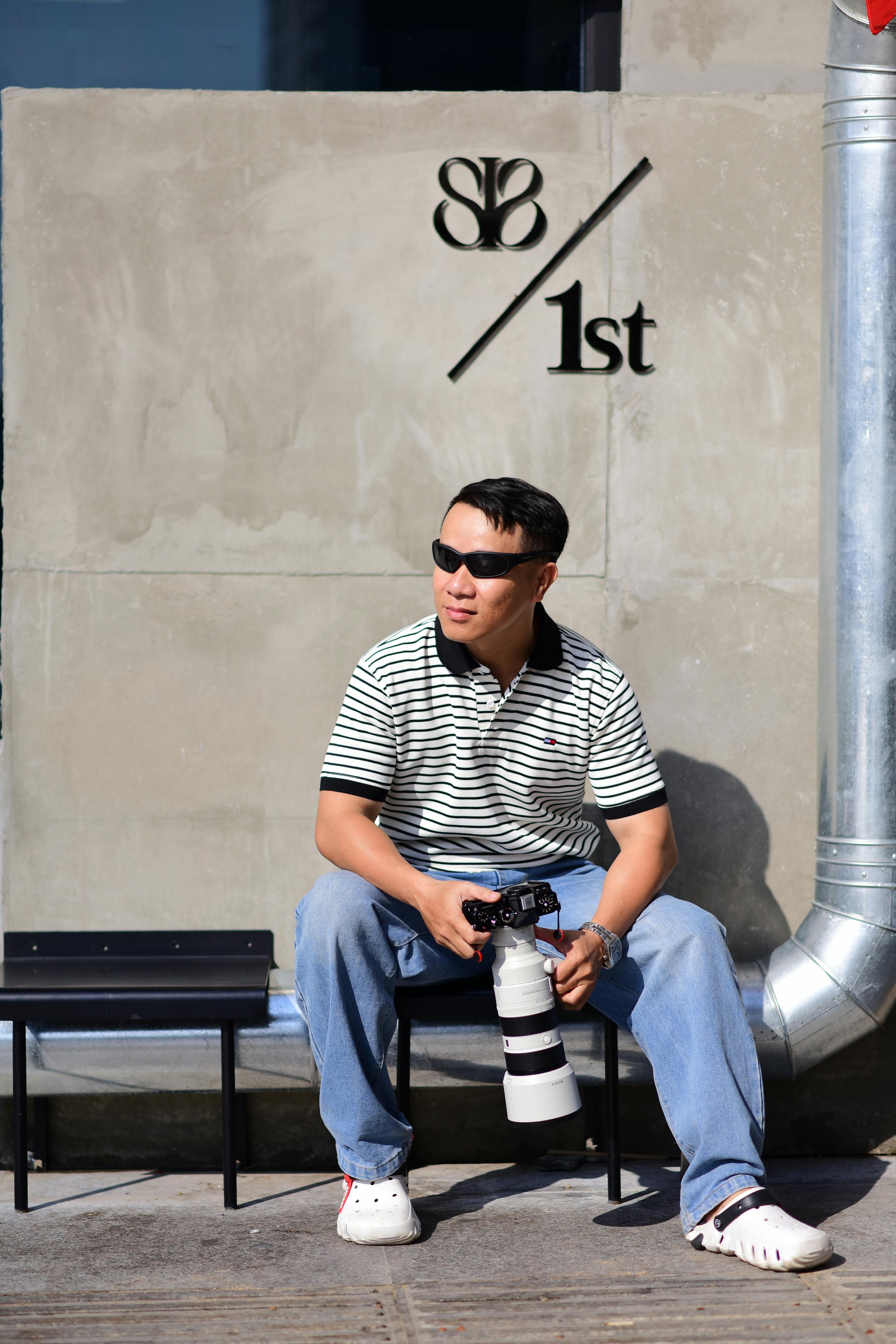 Man sitting on a bench with a camera, wearing sunglasses and casual clothes.