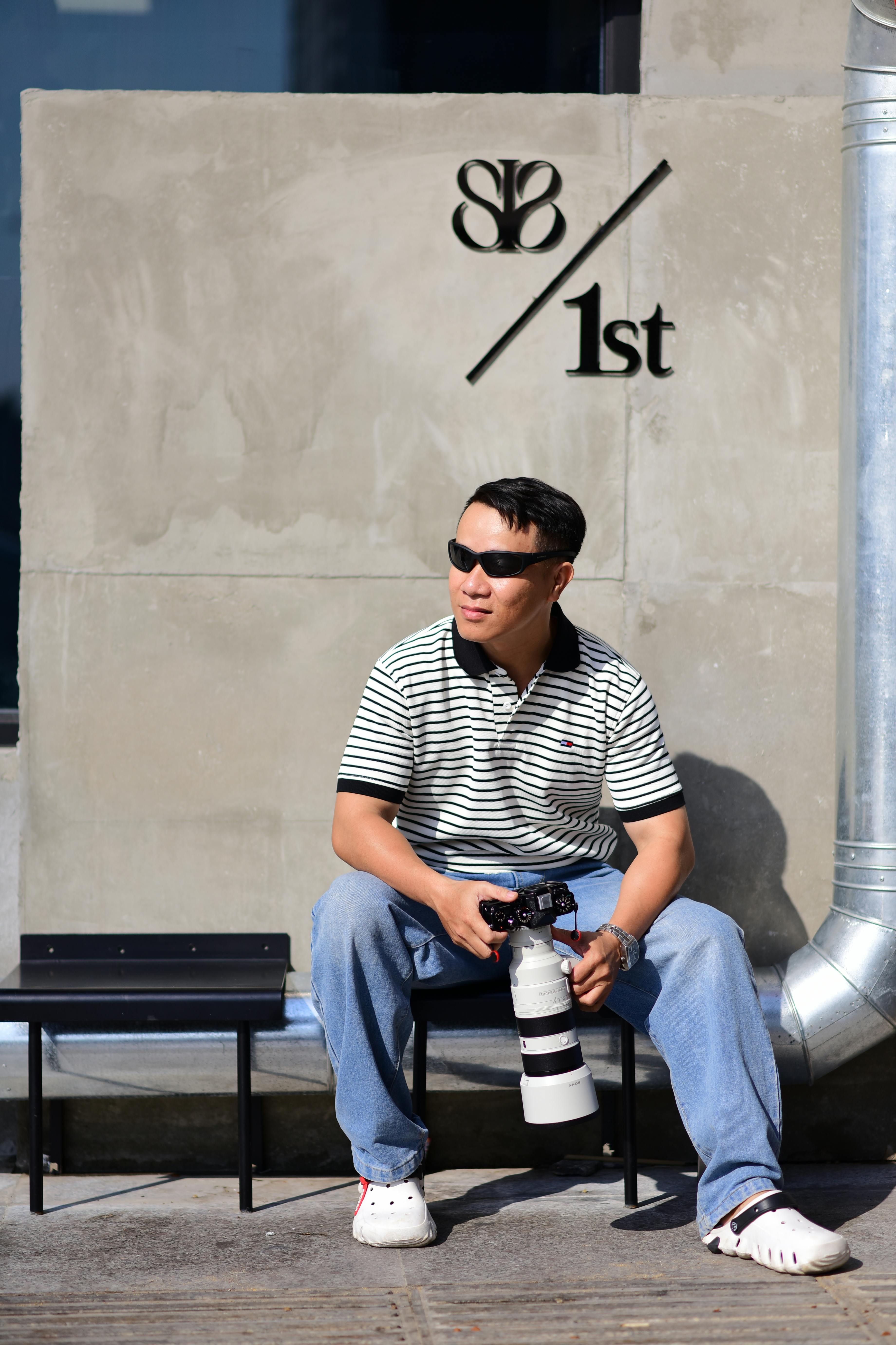 Δωρεάν Άνδρας φωτογράφος κάθεται με κάμερα, φορώντας γυαλιά ηλίου σε εξωτερικό χώρο, φόντο τσιμεντένιου τοίχου. Φωτογραφία από στοκ φωτογραφιών