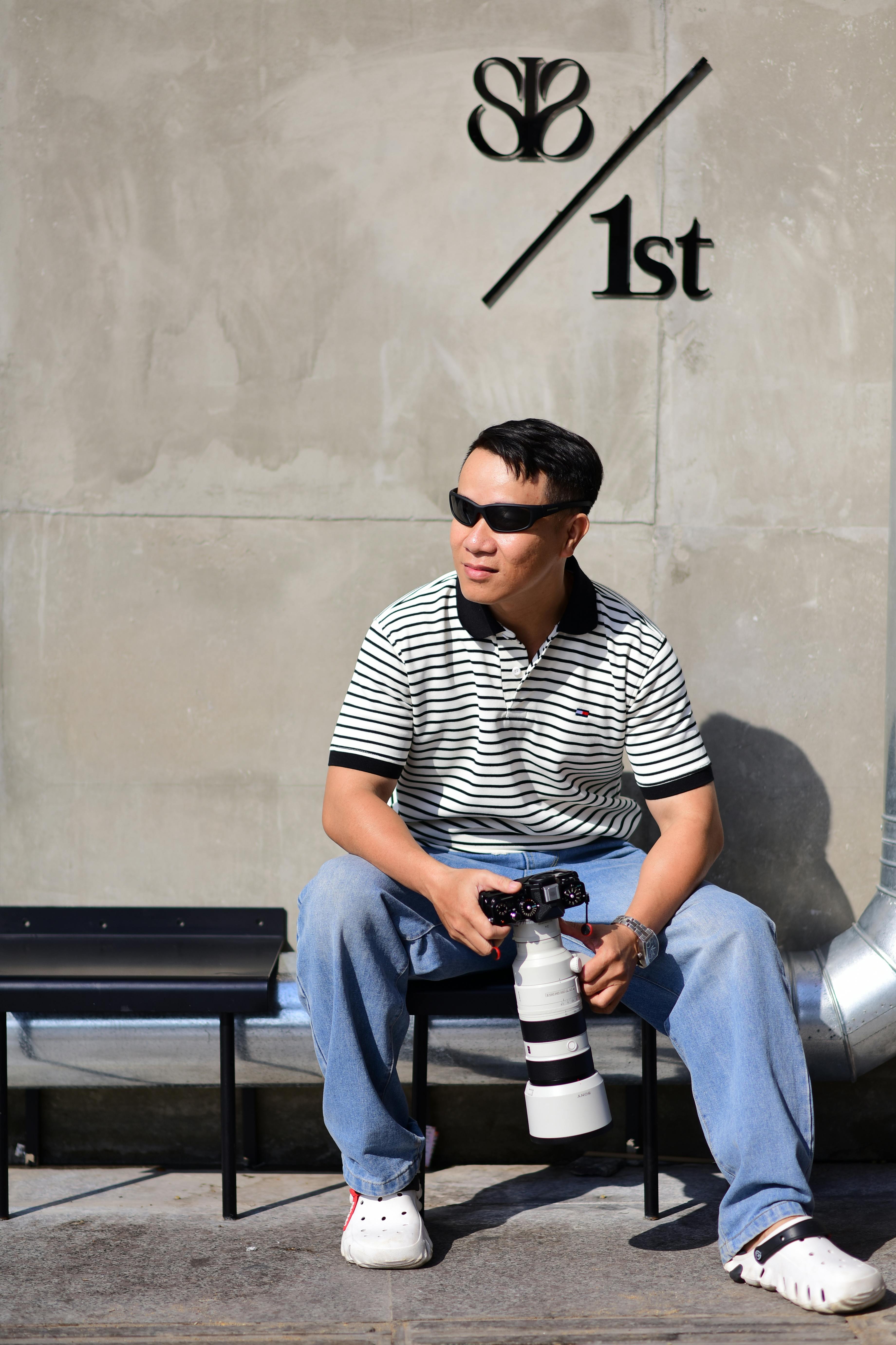 Man casually sits with camera, against a concrete urban backdrop, wearing sunglasses and striped shirt.