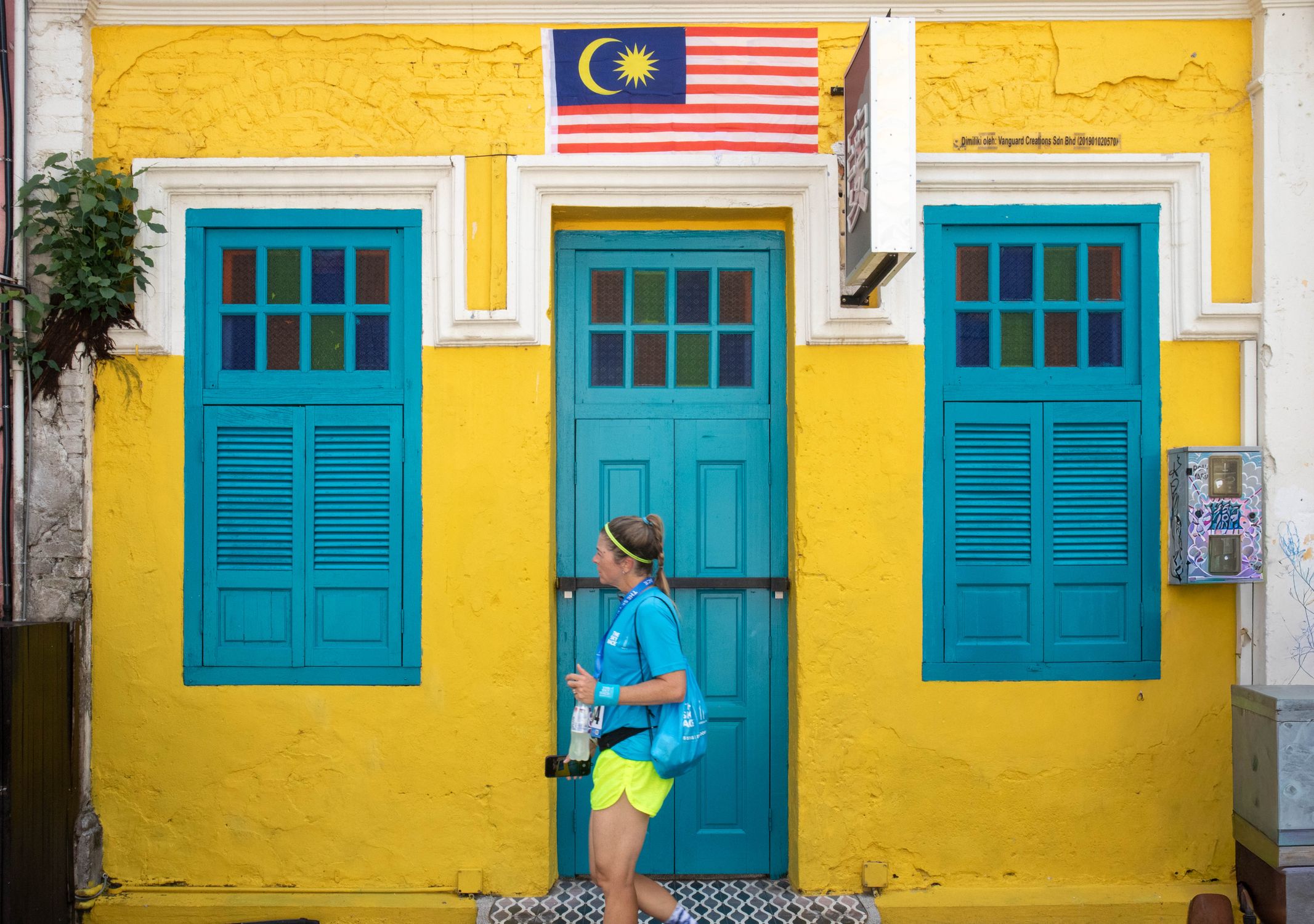A tourist walks by vibrant yellow and blue architecture adorned with a Malaysian flag.