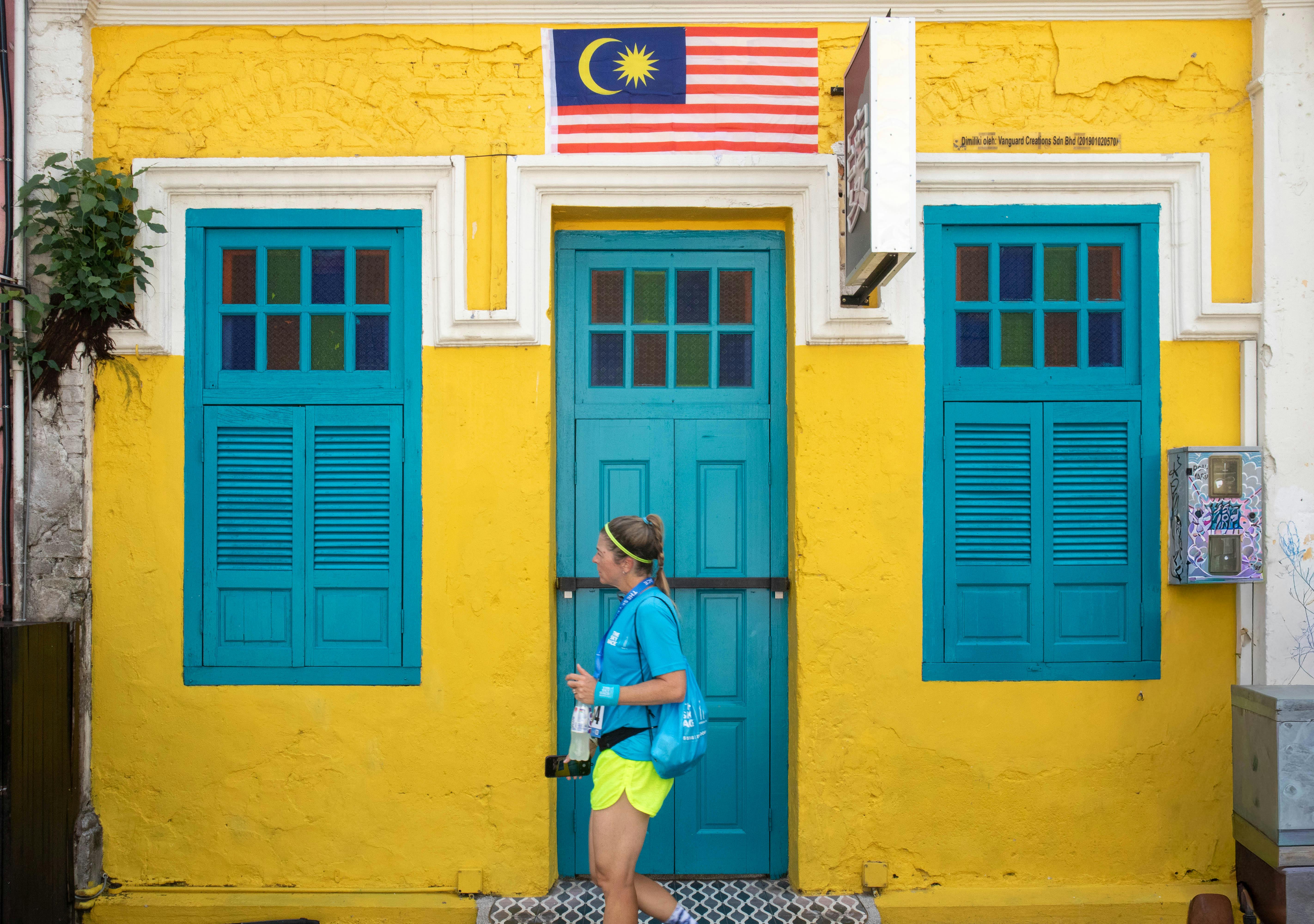 Kostenlos Ein Tourist spaziert an leuchtend gelb-blauen Gebäuden vorbei, die mit einer malaysischen Flagge geschmückt sind. Stock-Foto