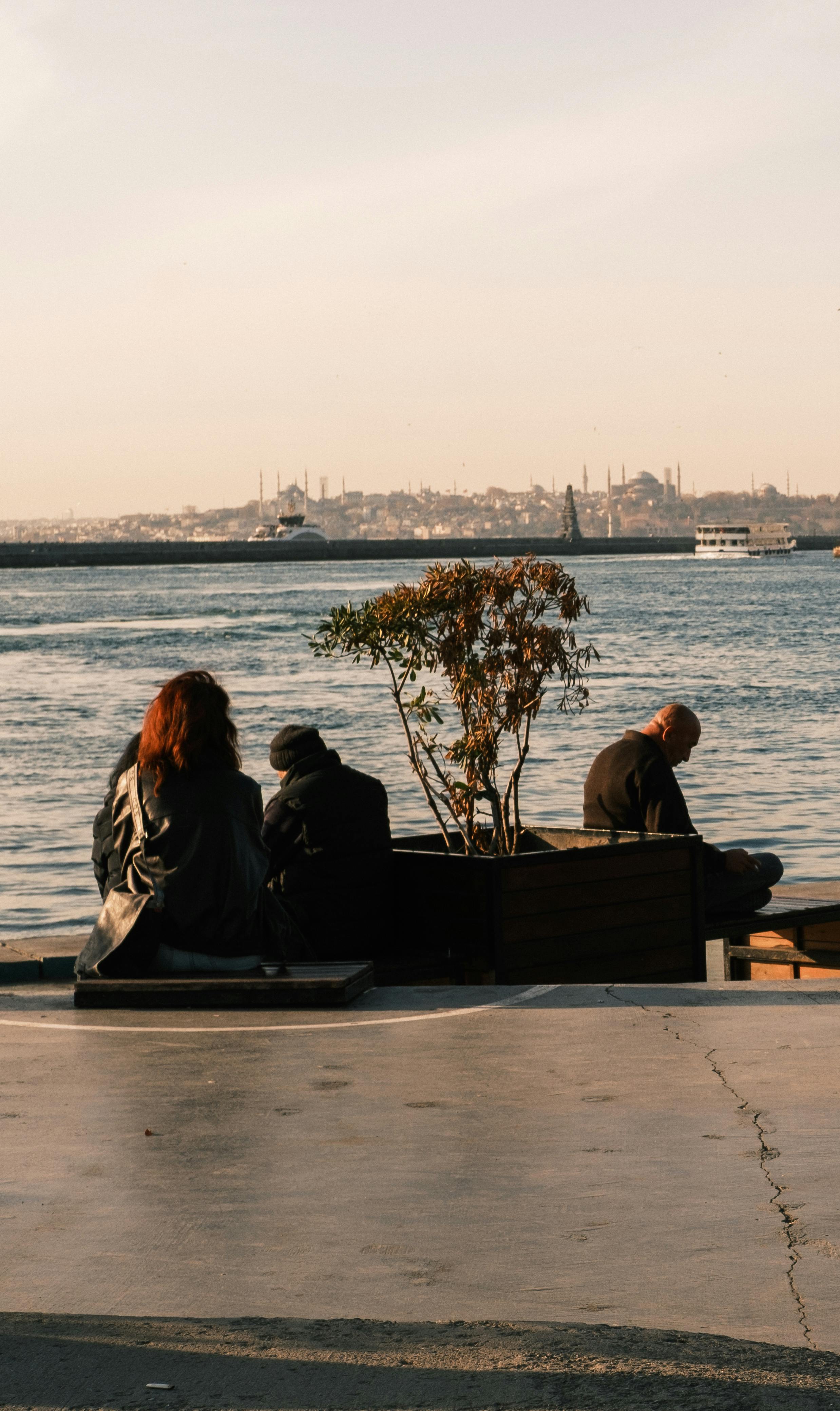 Kostenlos Menschen entspannen sich an einem Ufer und genießen einen ruhigen Sonnenuntergang mit Blick auf die Stadt in der Ferne. Stock-Foto