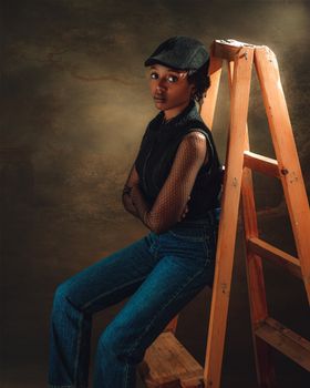 Stylish young woman in cap and denim posing indoors beside a wooden ladder.