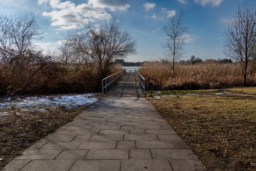 A tranquil winter pathway leading to a bridge over a lake under a partly cloudy sky.