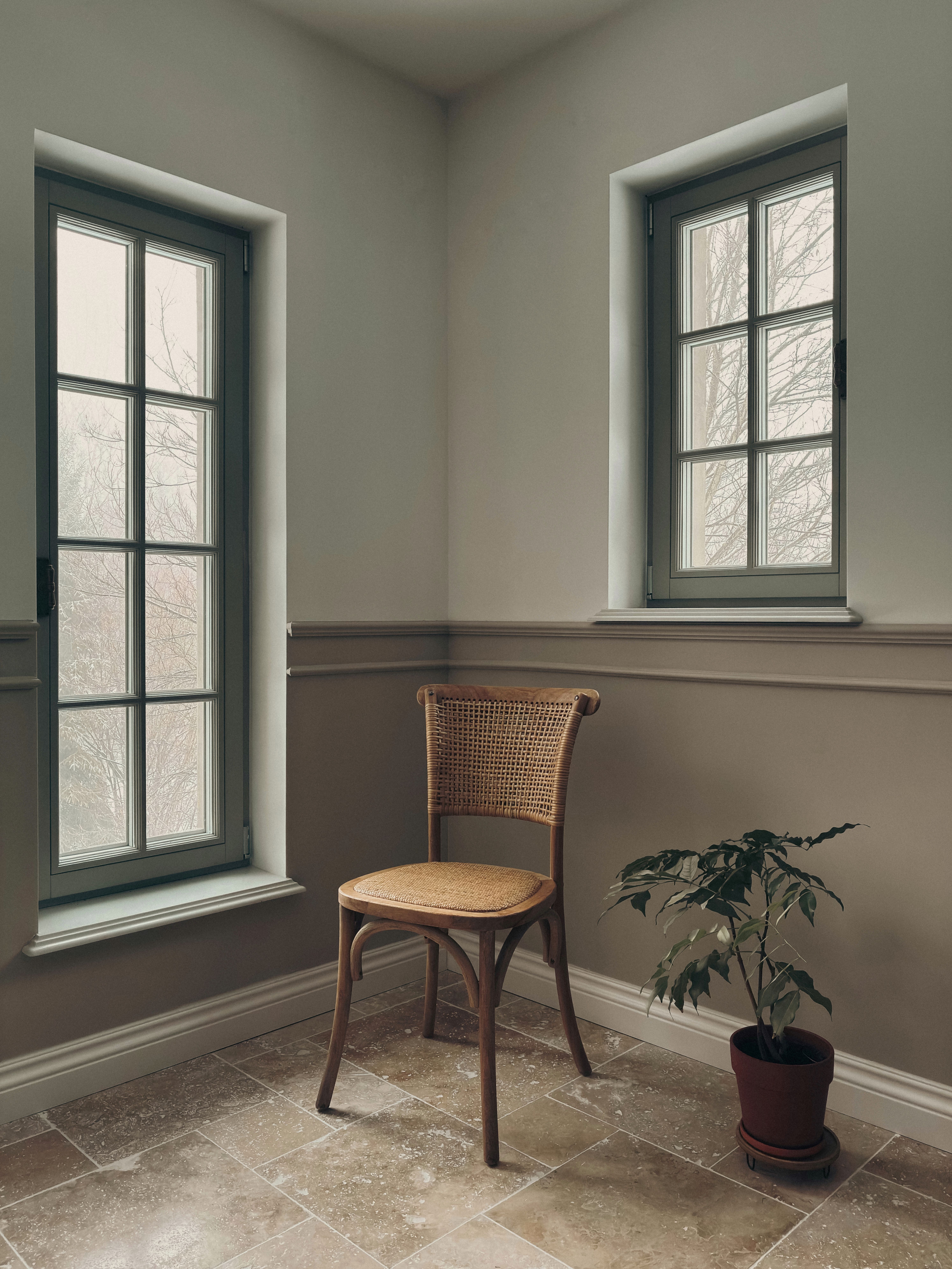 Serene indoor corner with a wooden chair and potted plant, near two windows.