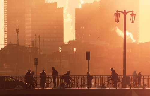 Free Silhouetted cyclists and pedestrians cross a city bridge at sunrise, with a skyline backdrop. Stock Photo
