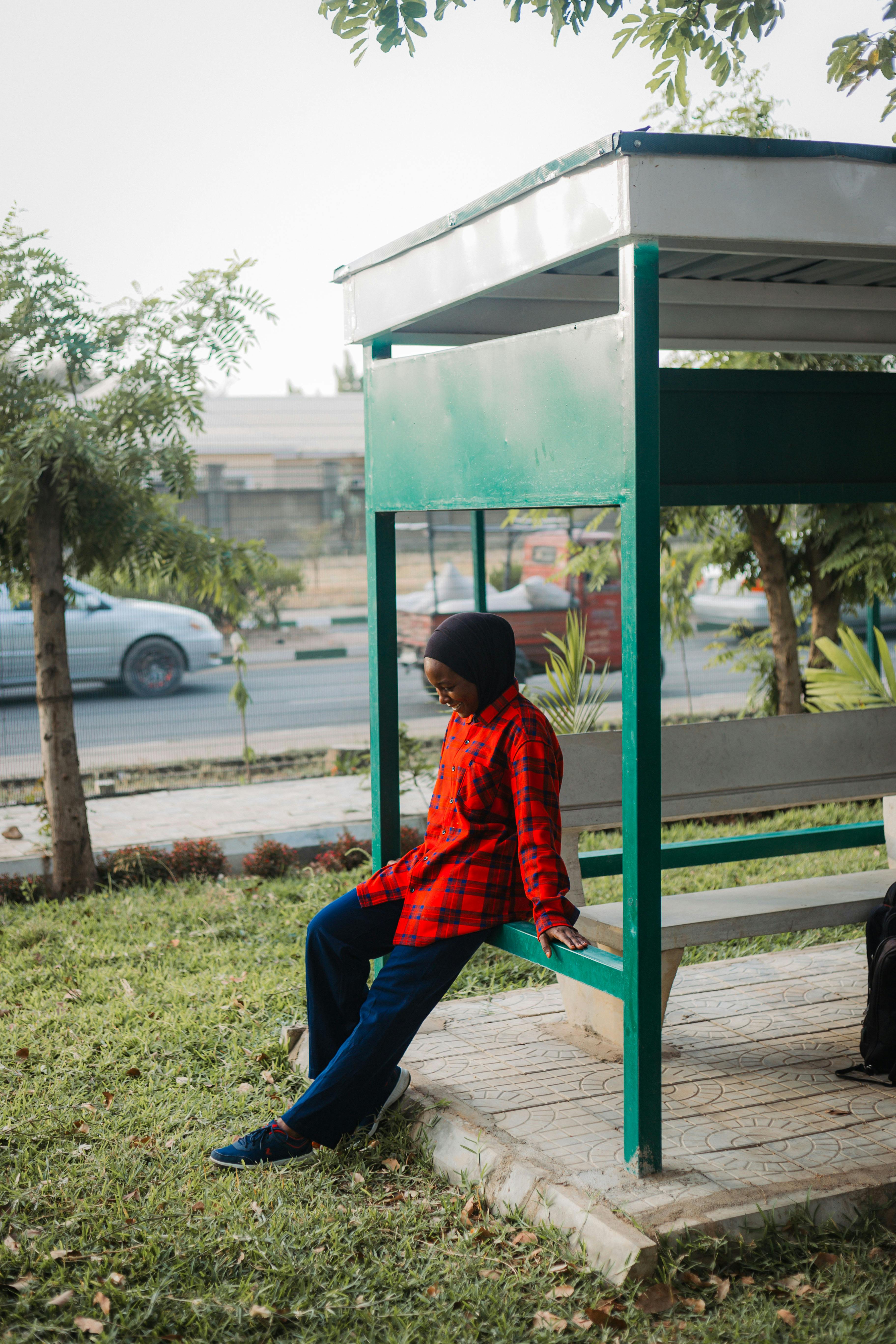 Kostenlos Junger Mann in rotem kariertem Hemd entspannt an einer Bushaltestelle im Freien in städtischer Umgebung. Stock-Foto