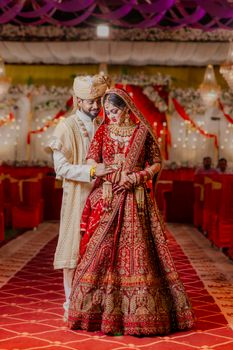 A beautifully dressed Indian couple in traditional wedding attire standing together indoors.