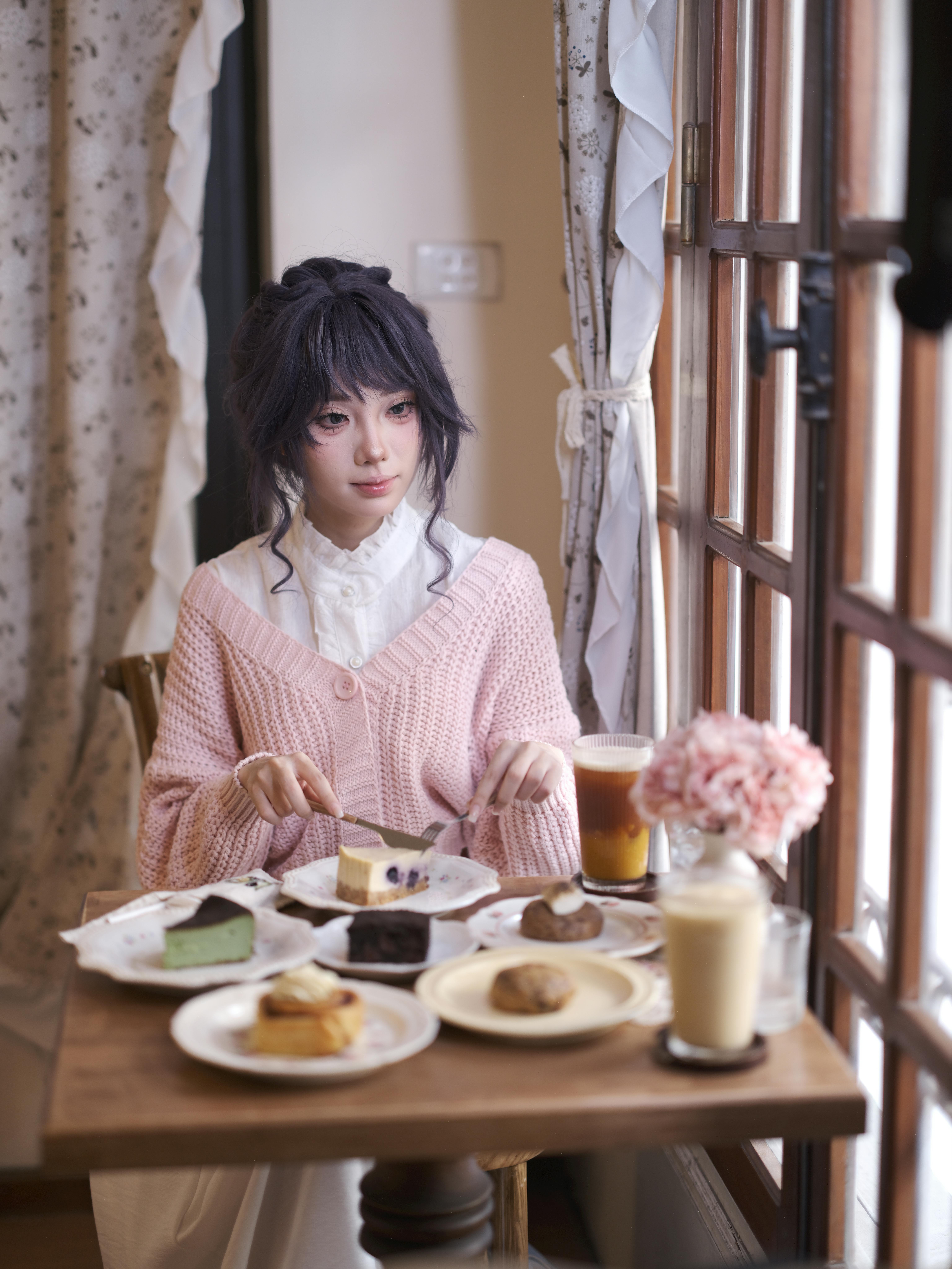 Woman enjoying desserts and drinks in a cozy cafe setting with natural light and floral ambiance.