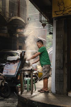 Man cooking from street food stall in urban alley, steam rising.