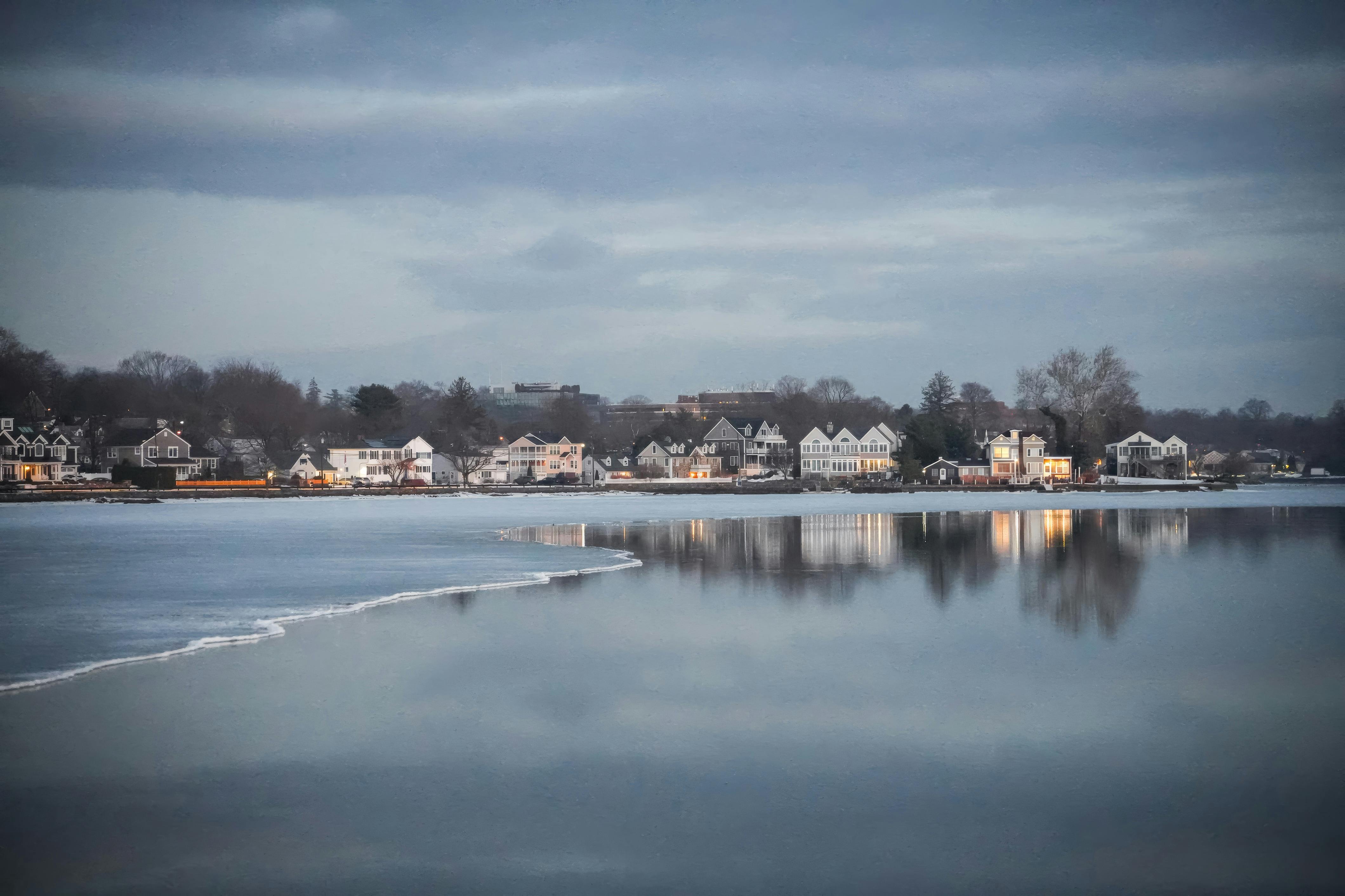 Kostenlos Friedlicher Winterabend an einem zugefrorenen See mit gemütlichen Häusern und Spiegelungen. Stock-Foto