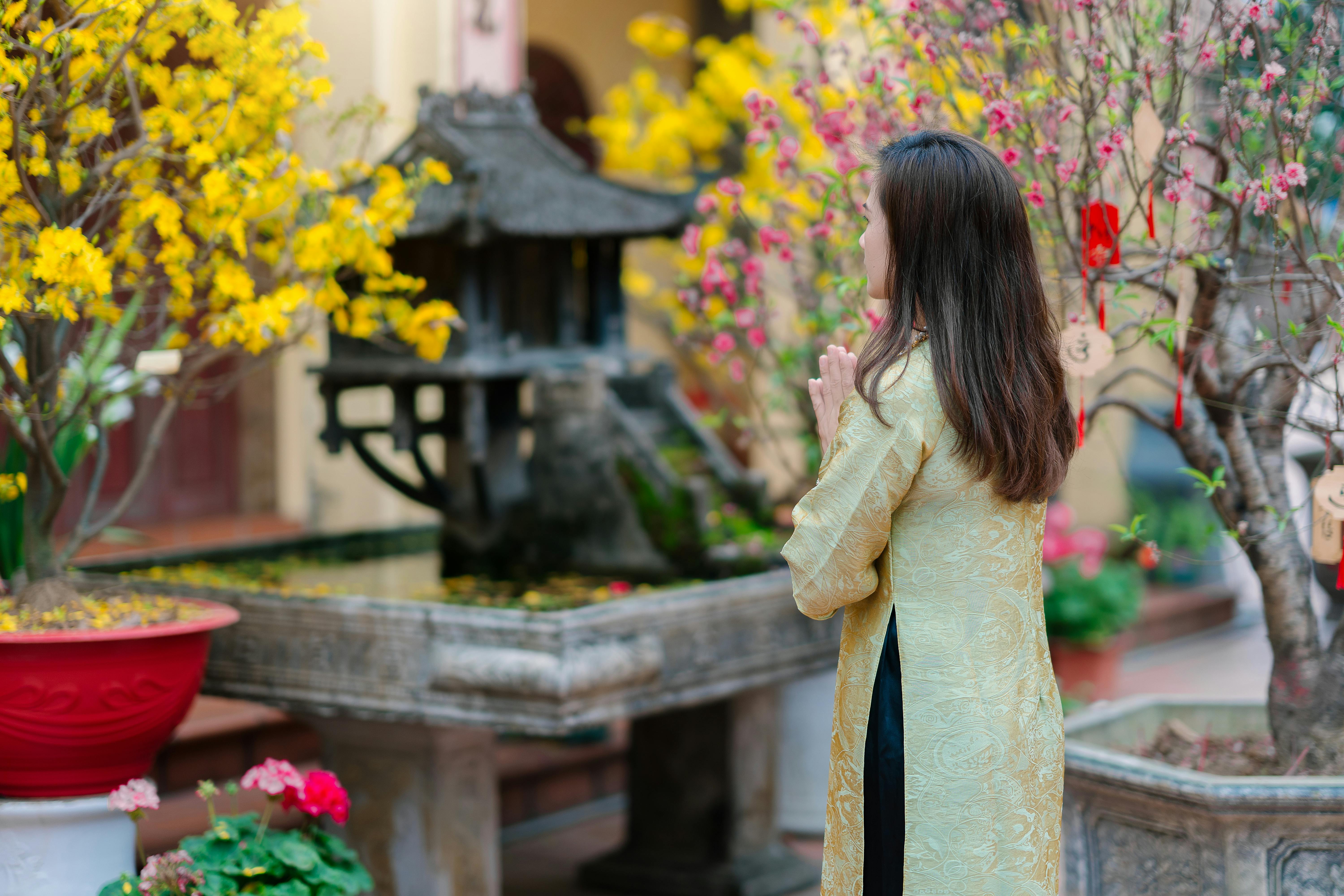 Kostenlos Eine Frau in traditioneller Kleidung betet neben farbenprächtigen blühenden Bäumen in einem Tempel. Stock-Foto